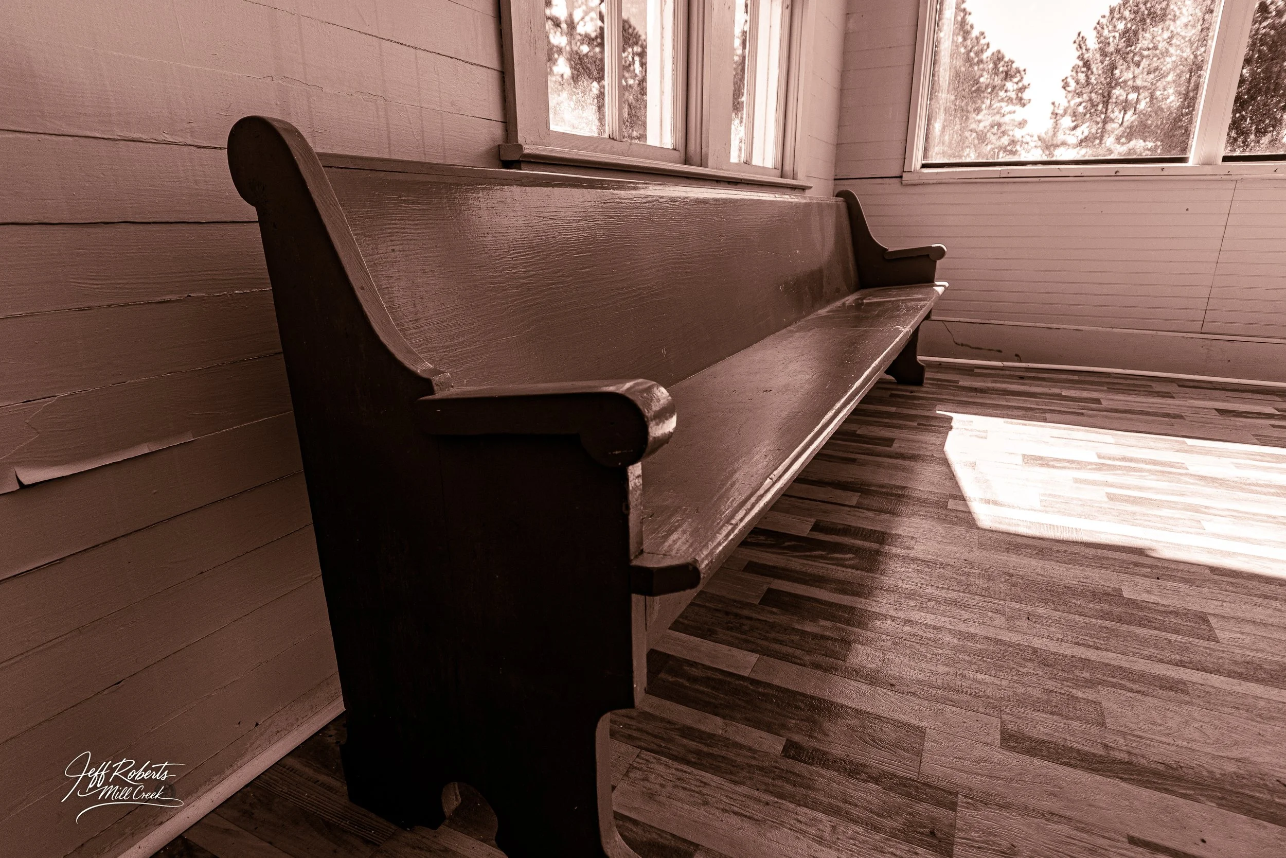 An old wooden church pew inside a sunlit room with windows, wooden floor, and light-colored paneled walls.