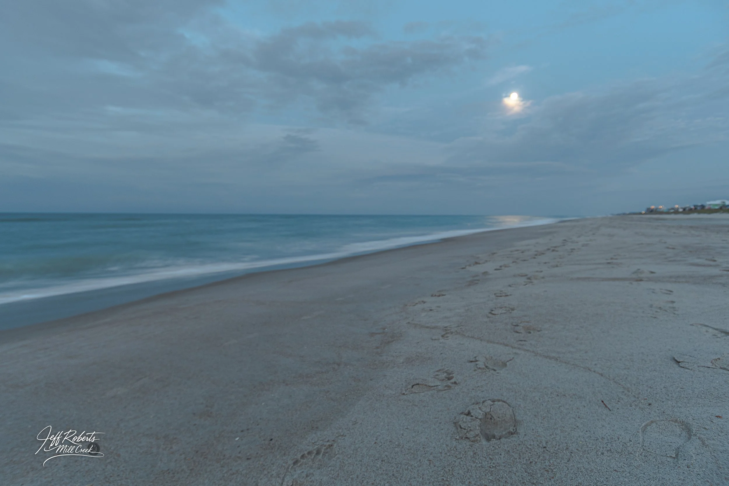 Beach scene at dusk with footprints in the sand, calm ocean waves, cloudy sky, and moon visible