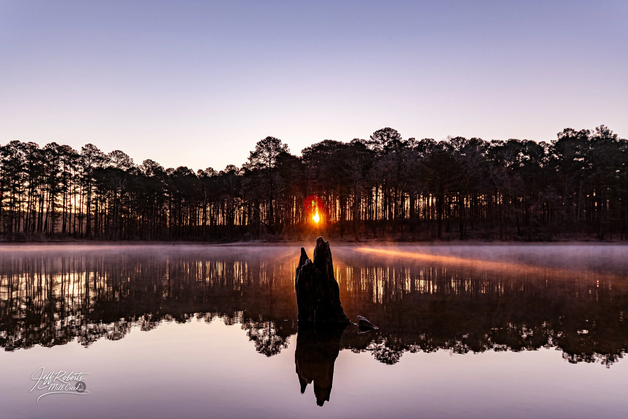 Sunset over a calm lake with a reflection of trees and a tree stump in the water.