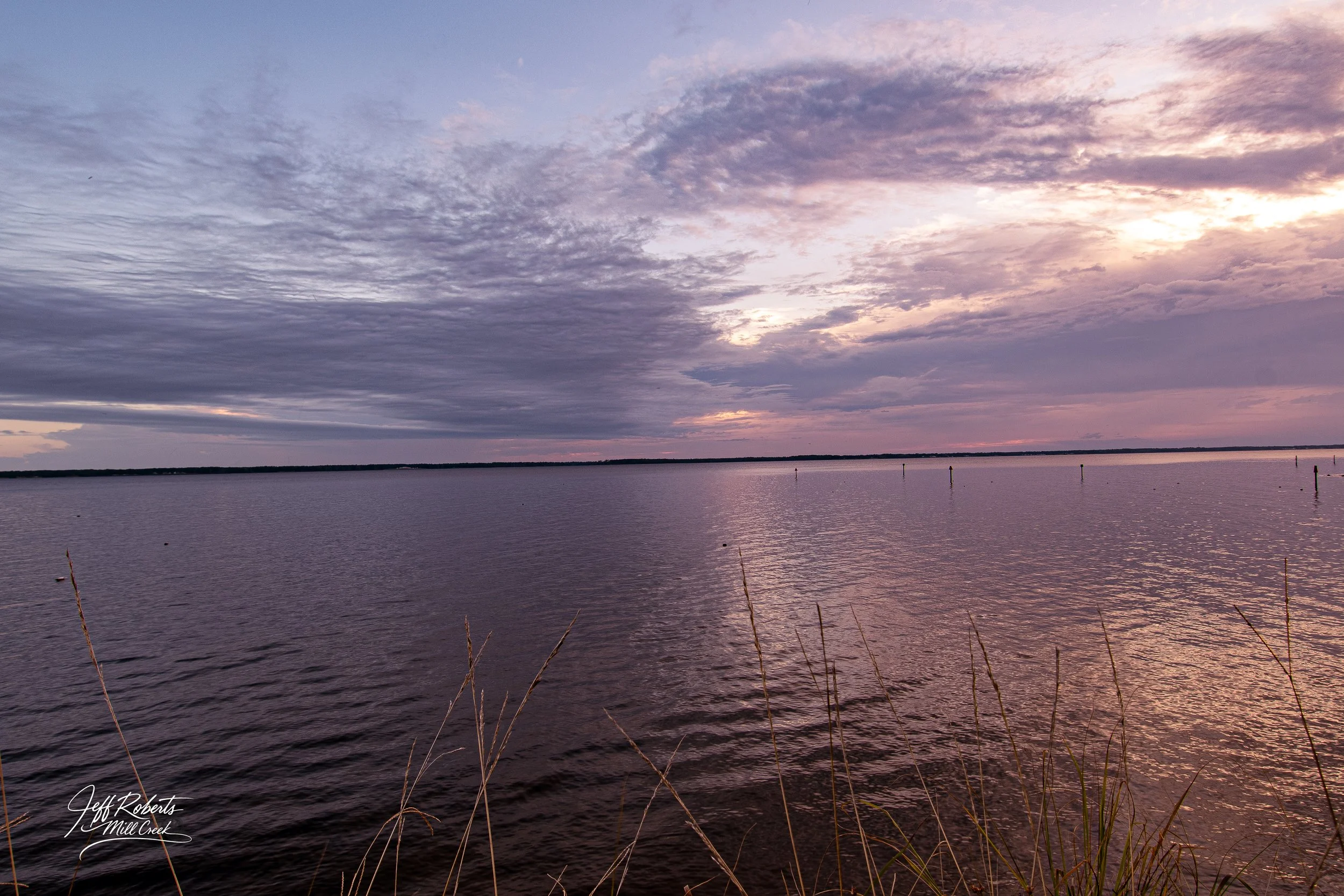 Calm body of water reflecting a colorful, cloudy sky at dusk or dawn with a distant shoreline and some grass in the foreground.