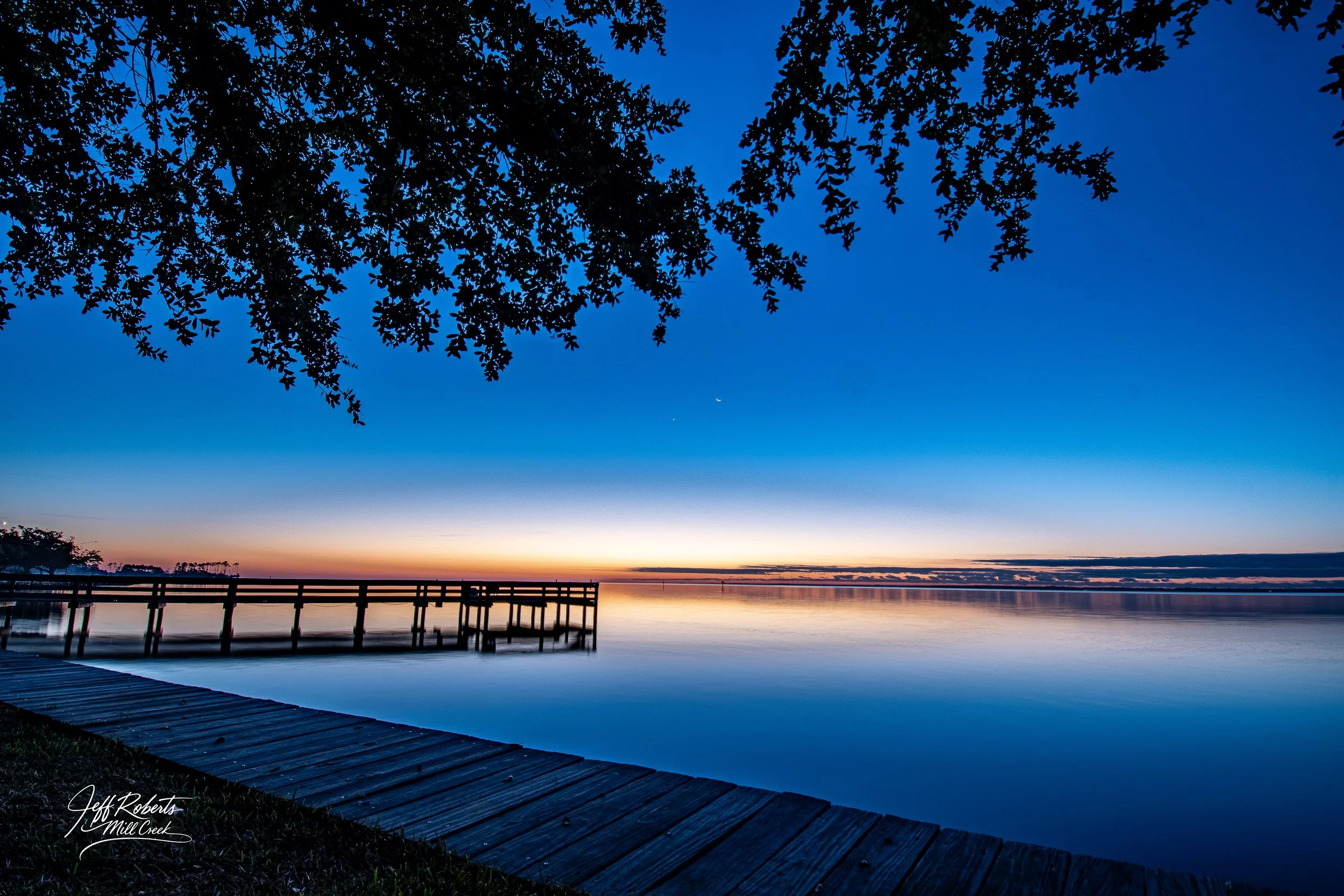Sunset over a calm lake with a wooden pier, under a tree with overhanging branches.