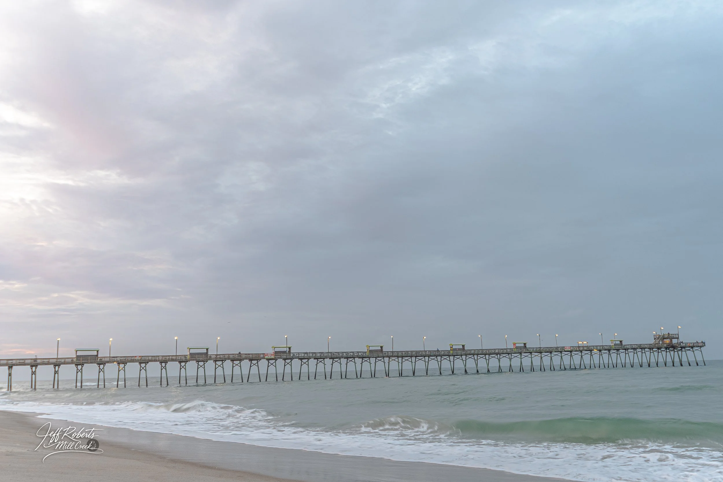 Long pier extending into the ocean with small structures and lights, cloudy sky overhead, waves hitting the sandy beach in the foreground.