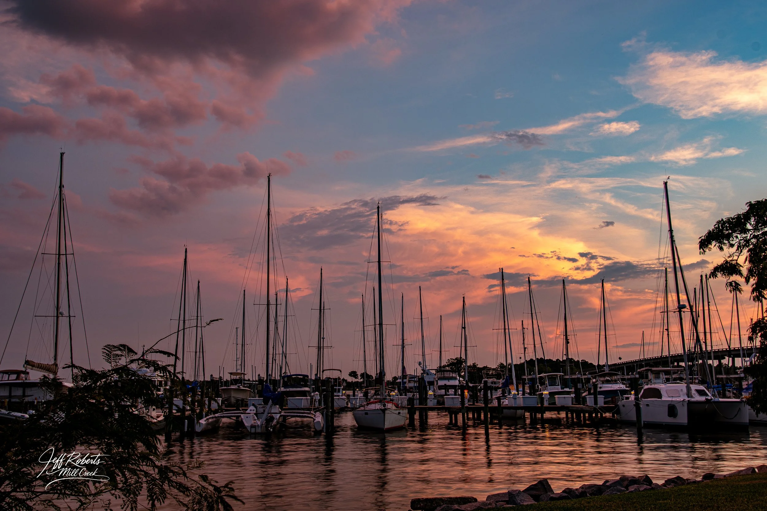 A marina at sunset with sailboats docked, reflecting in the water, with pink, purple, and blue clouds in the sky.