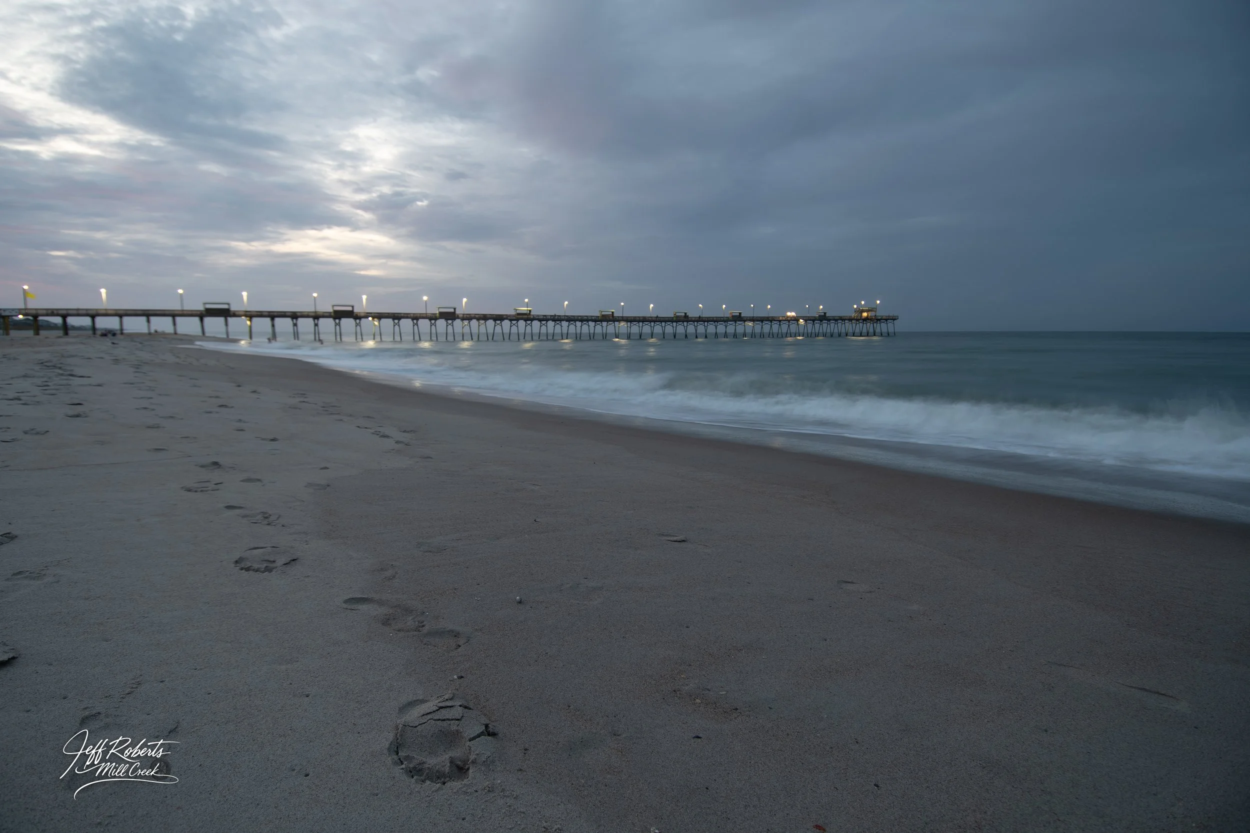 Footprints in the sand on a beach near a pier extending into the ocean during cloudy weather, with soft waves and overcast sky.
