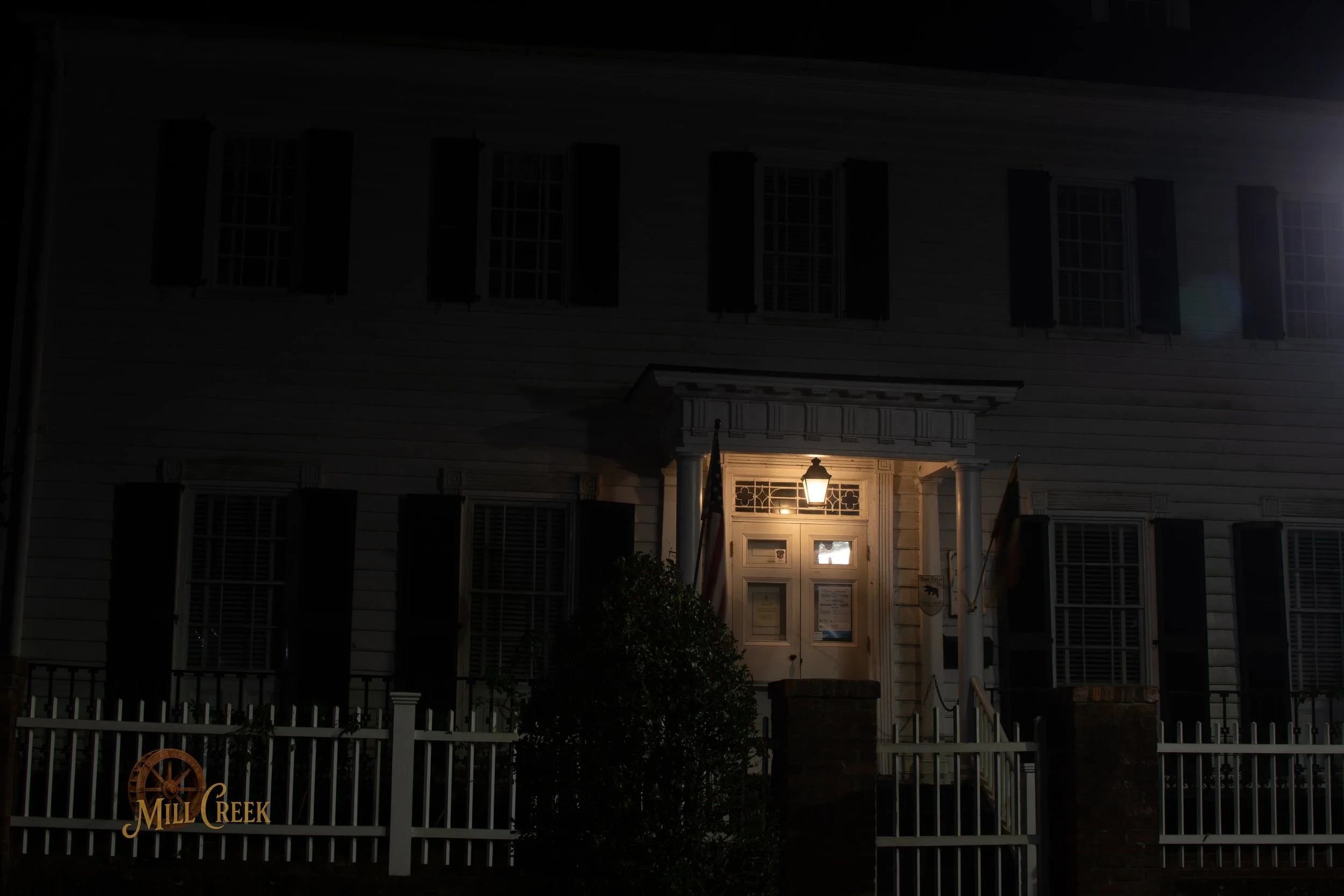 A dark, nighttime view of an old, white, two-story building with black shutters. A lit lantern is above the front door, illuminating the entrance. There is a white picket fence with a sign that says 'Mill Creek' along the fence in the front yard.