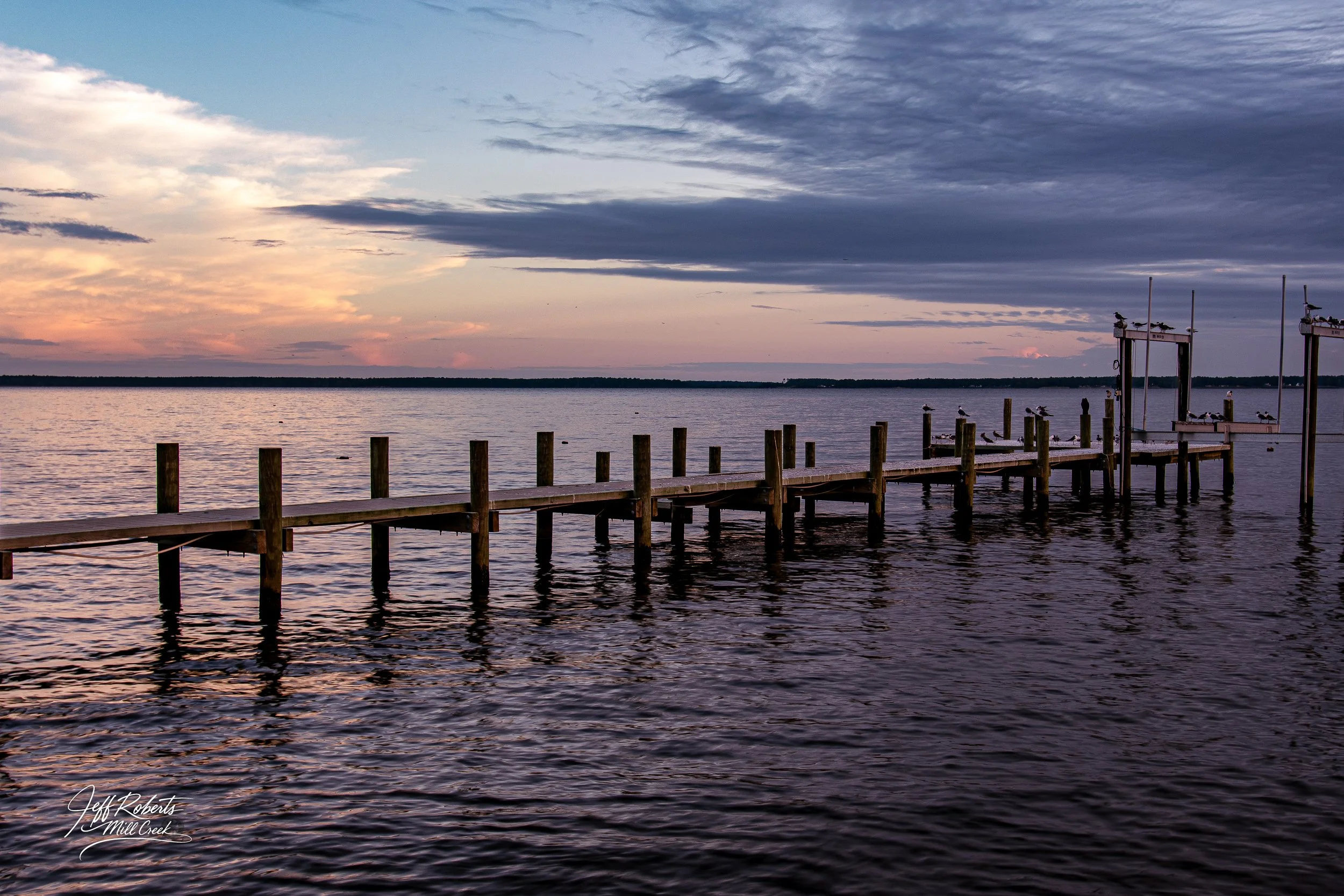 A wooden dock extending into a calm body of water during sunset, with a partly cloudy sky.