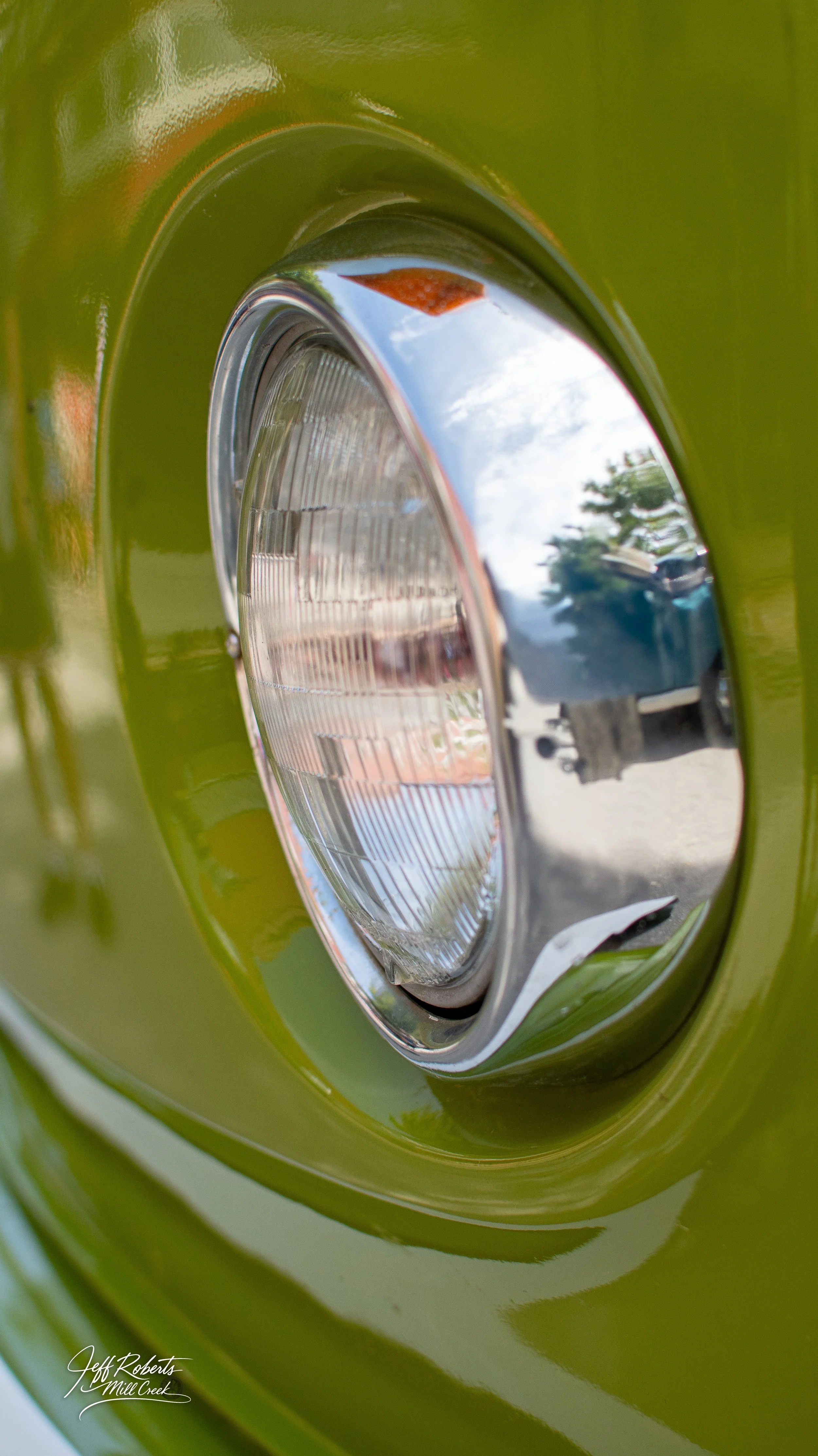 Close-up of a vintage green car headlight with reflective chrome detail, showing the sky and trees reflected in the shiny surface.