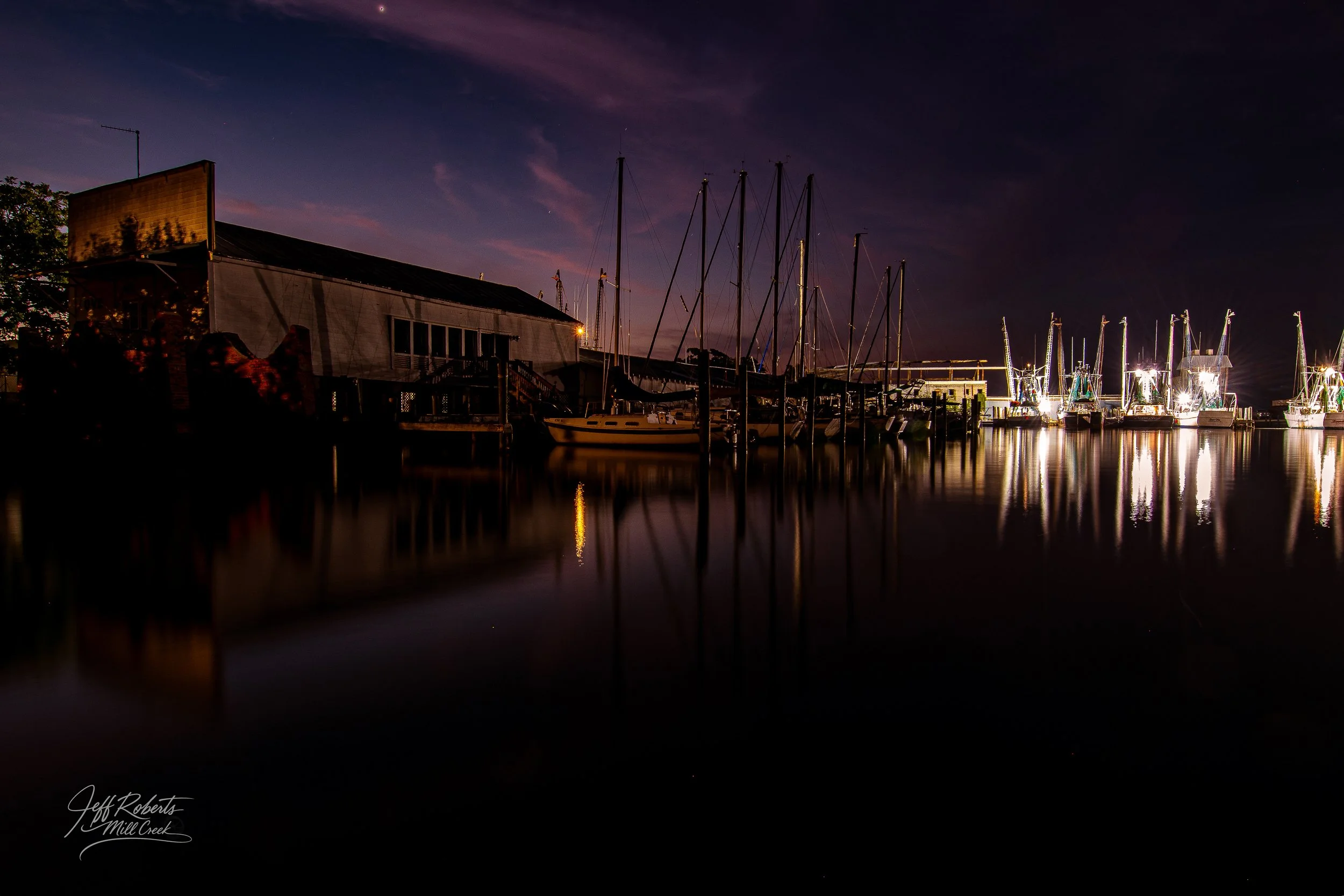 Nighttime view of a marina with sailboats and yachts docked along the water, illuminated by bright lights on the right, with reflections on the water, and a building with a dark sky above.