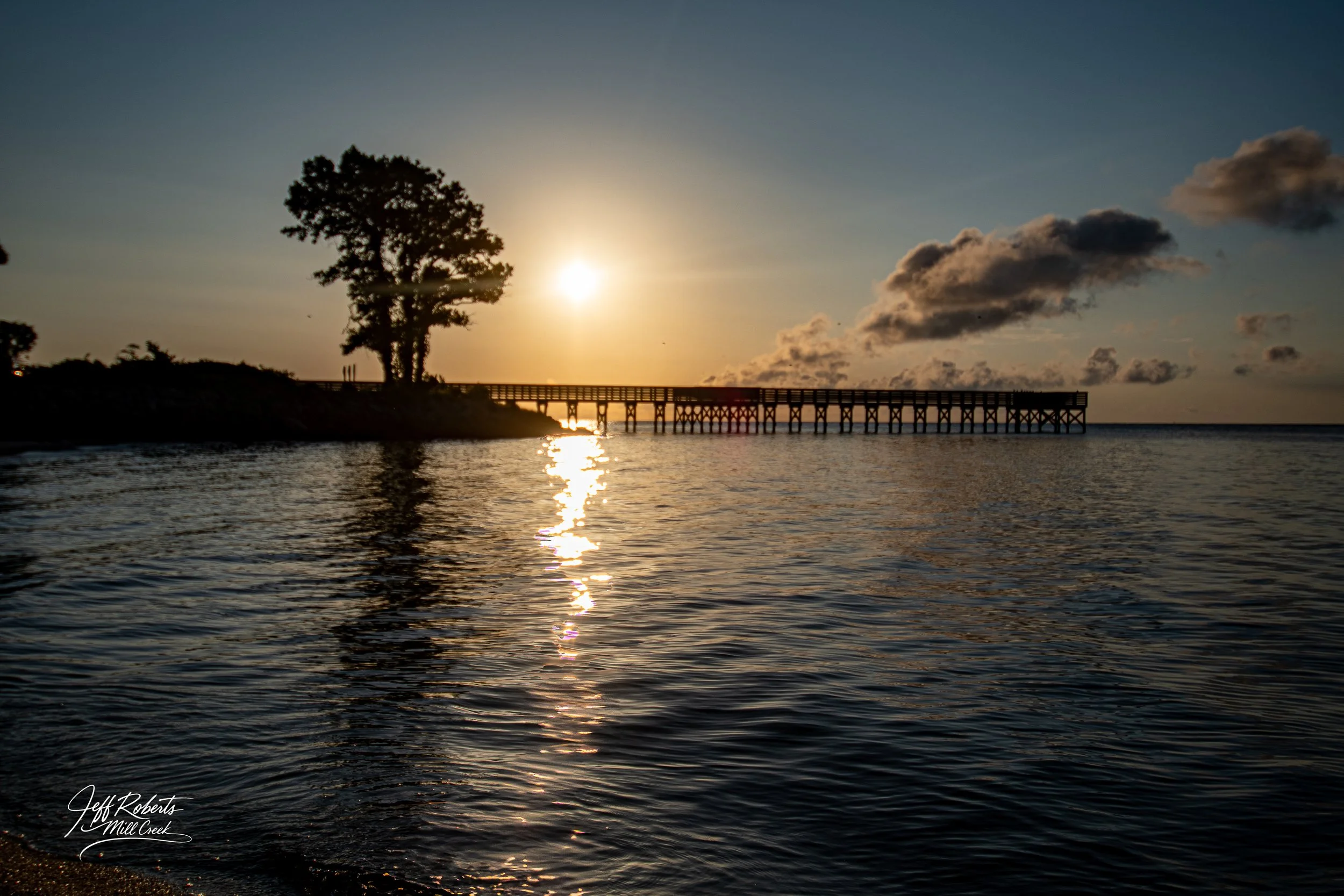 Sunset over the water with a wooden pier extending into the ocean, silhouette of trees on the land, and partly cloudy sky.
