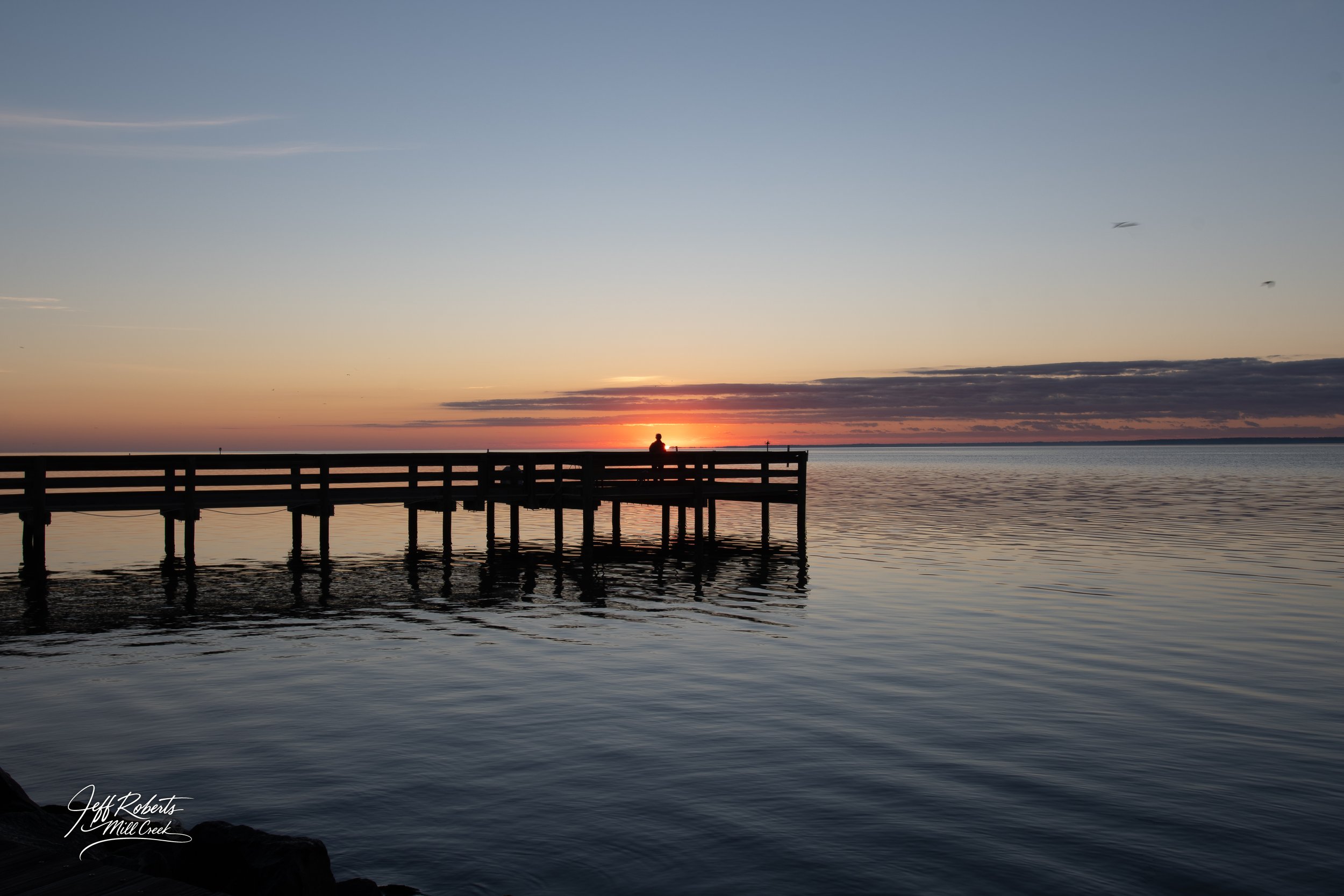 Sunset view over a calm body of water with a wooden pier extending into the water and a person sitting at the end of the pier, silhouettes against the colorful sky.