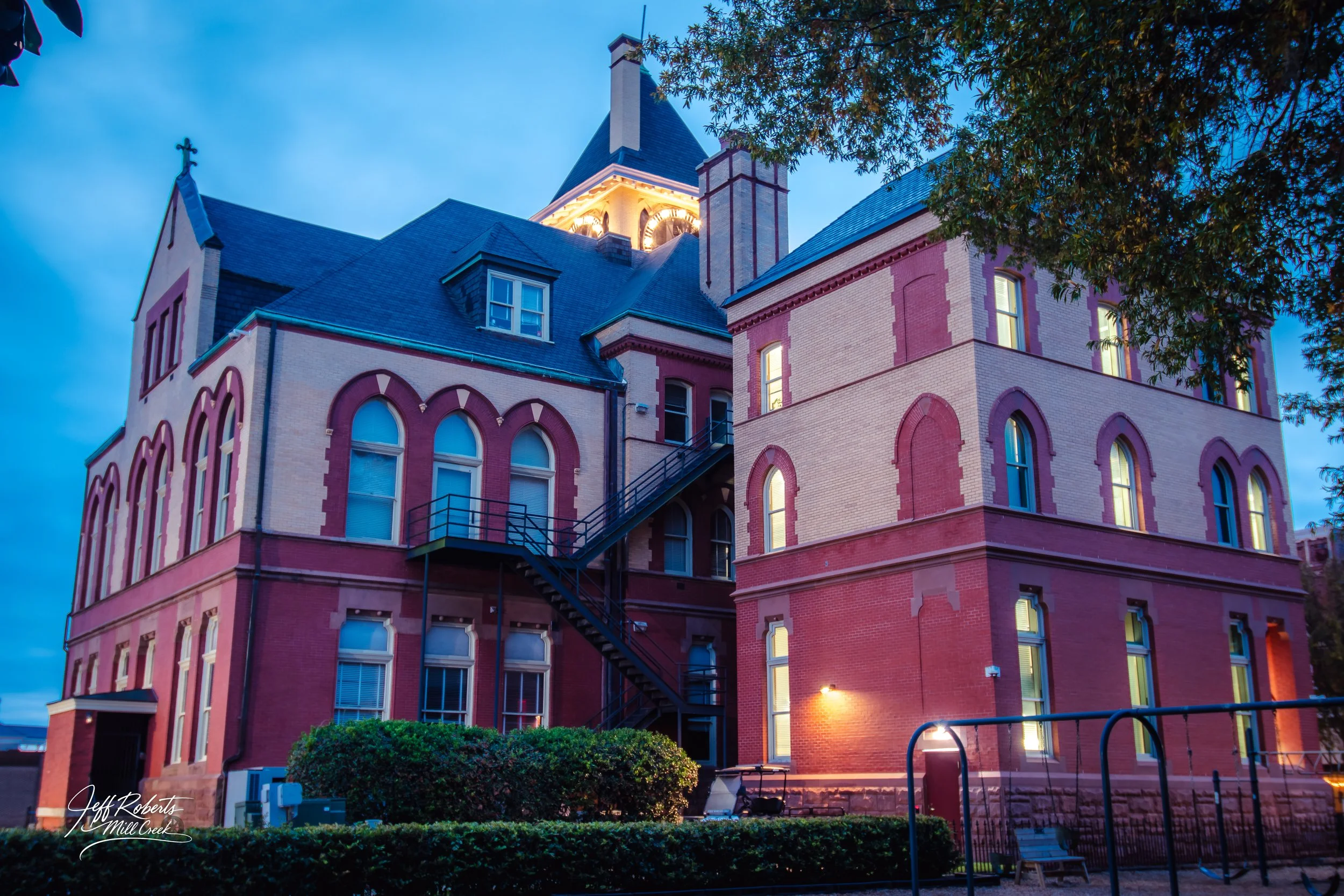Historic brick building illuminated at dusk with arched windows and a clock tower, surrounded by trees and a small garden.