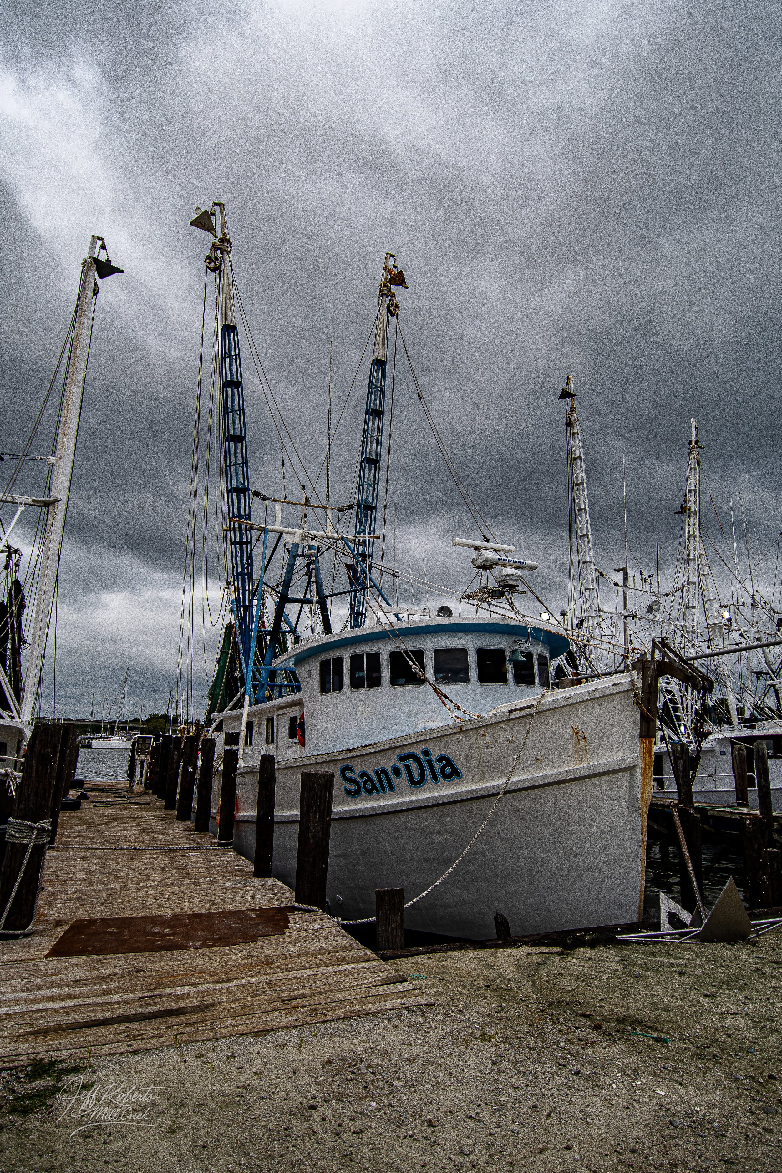 A marina with boats docked on a cloudy day, featuring a boat named San Daa in the foreground and dark stormy skies.