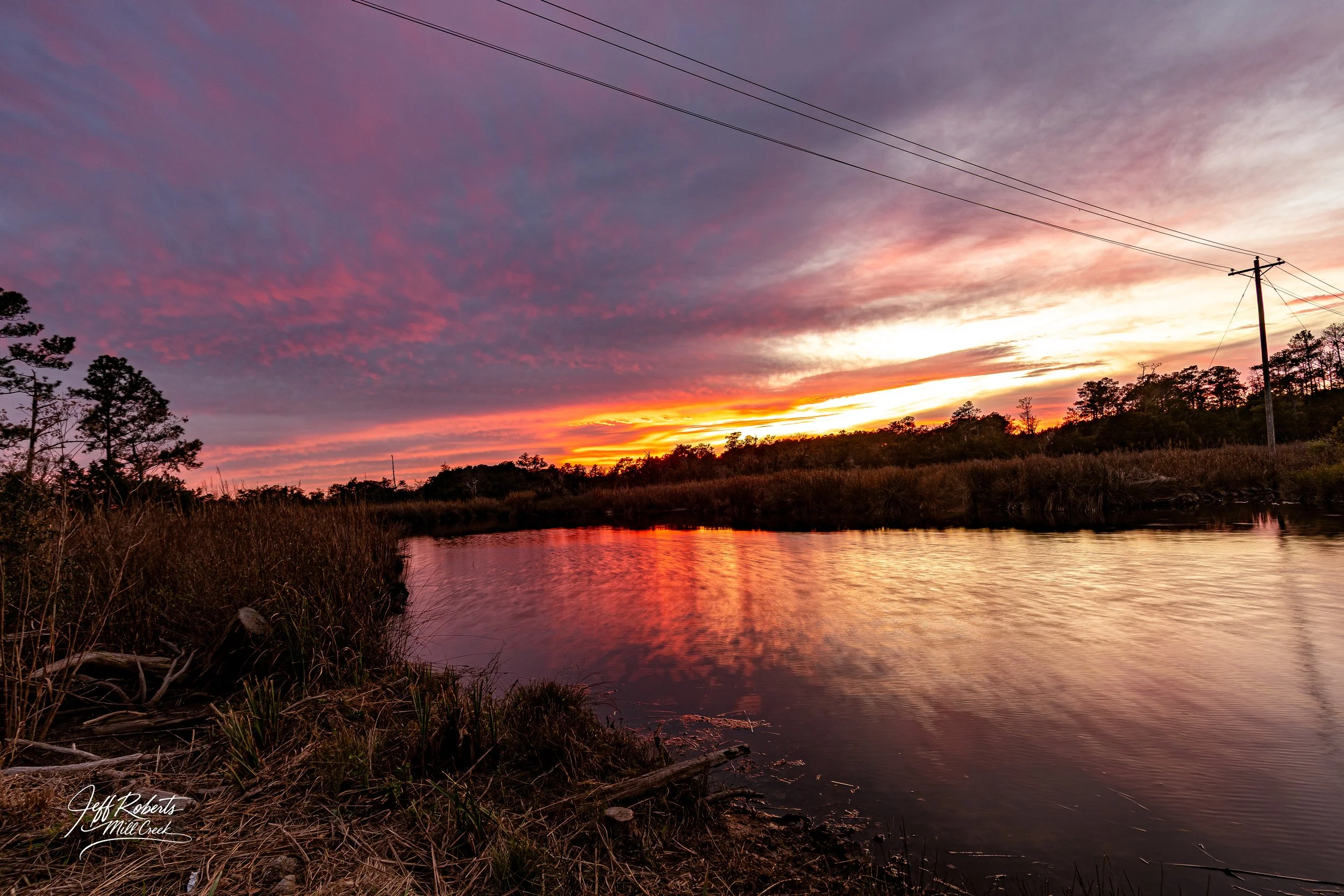 Sunset over a river with colorful clouds and silhouettes of trees and power lines.