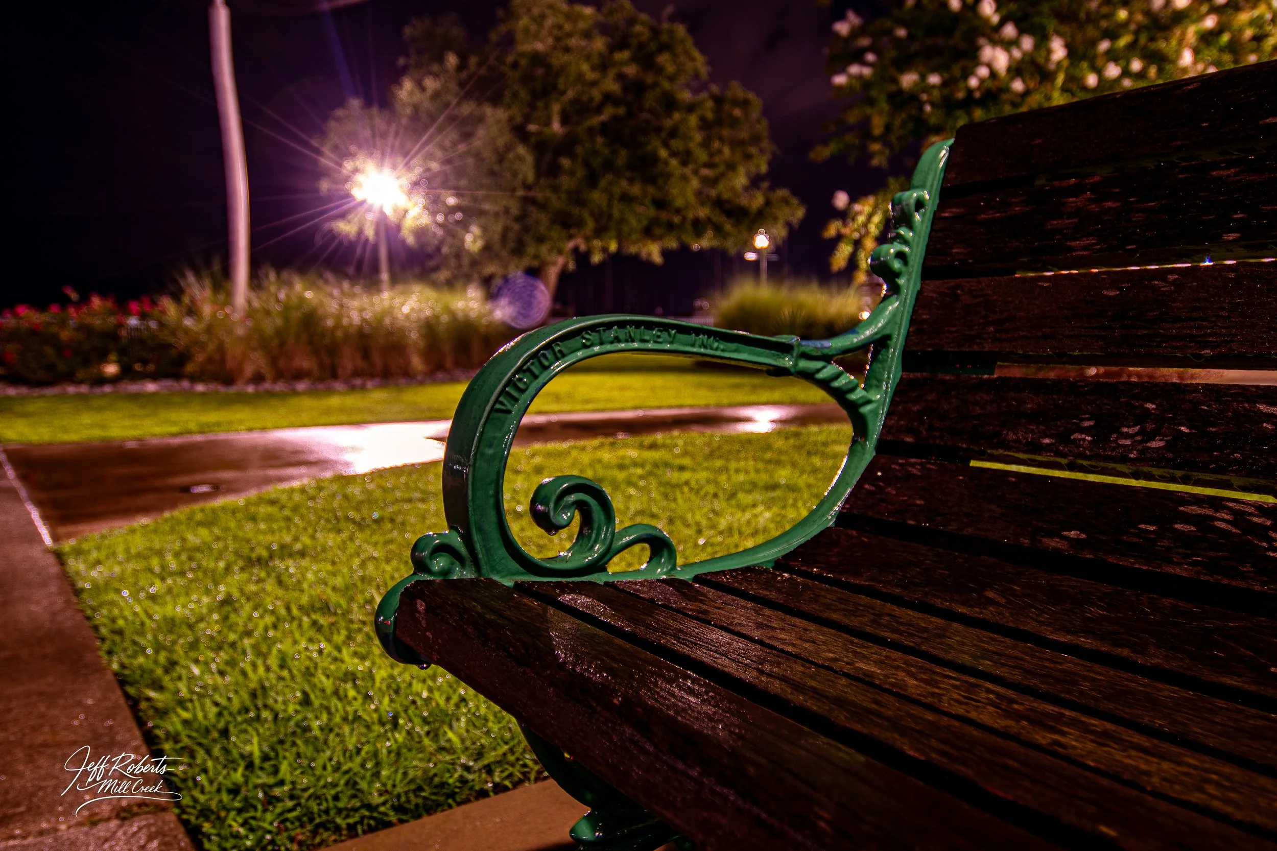 Nighttime photo of a park bench with green metal armrest and weathered wooden planks, illuminated by a street lamp with a blurred background of trees and flowers.