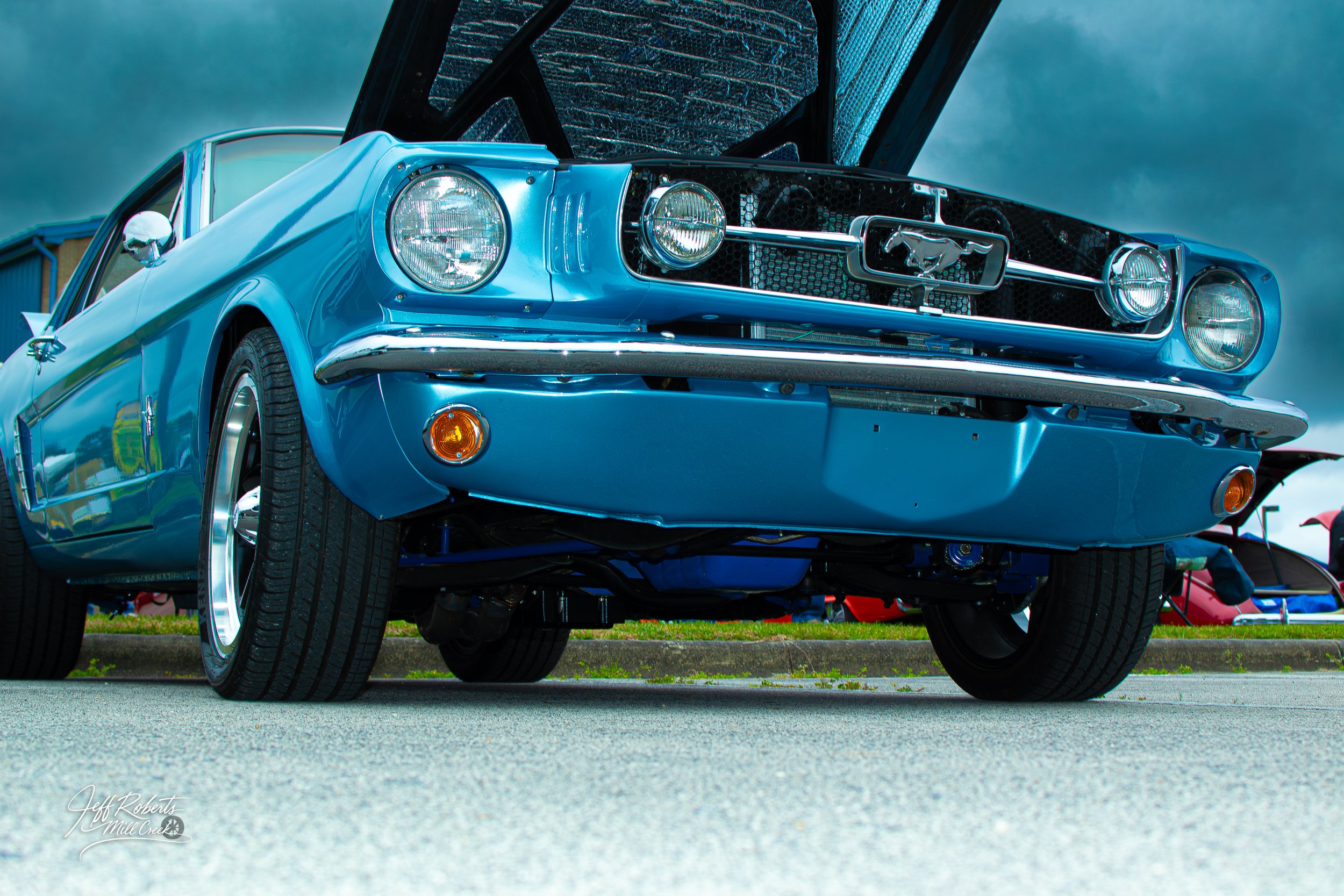 A classic blue Ford Mustang car with its hood open, on display at a car show.