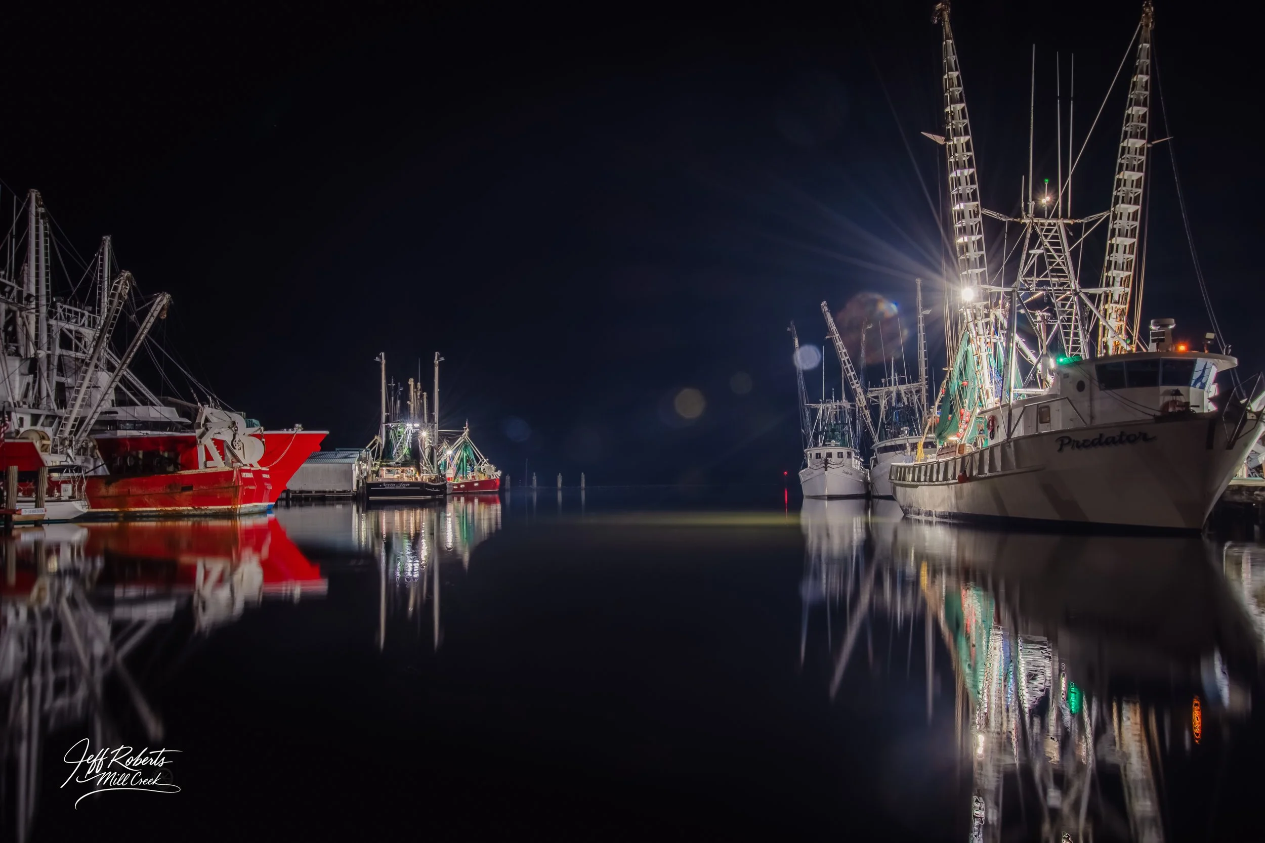 Nighttime view of fishing boats docked at a marina, with their reflections visible on the calm water, and bright lights illuminating the boats against the dark sky.