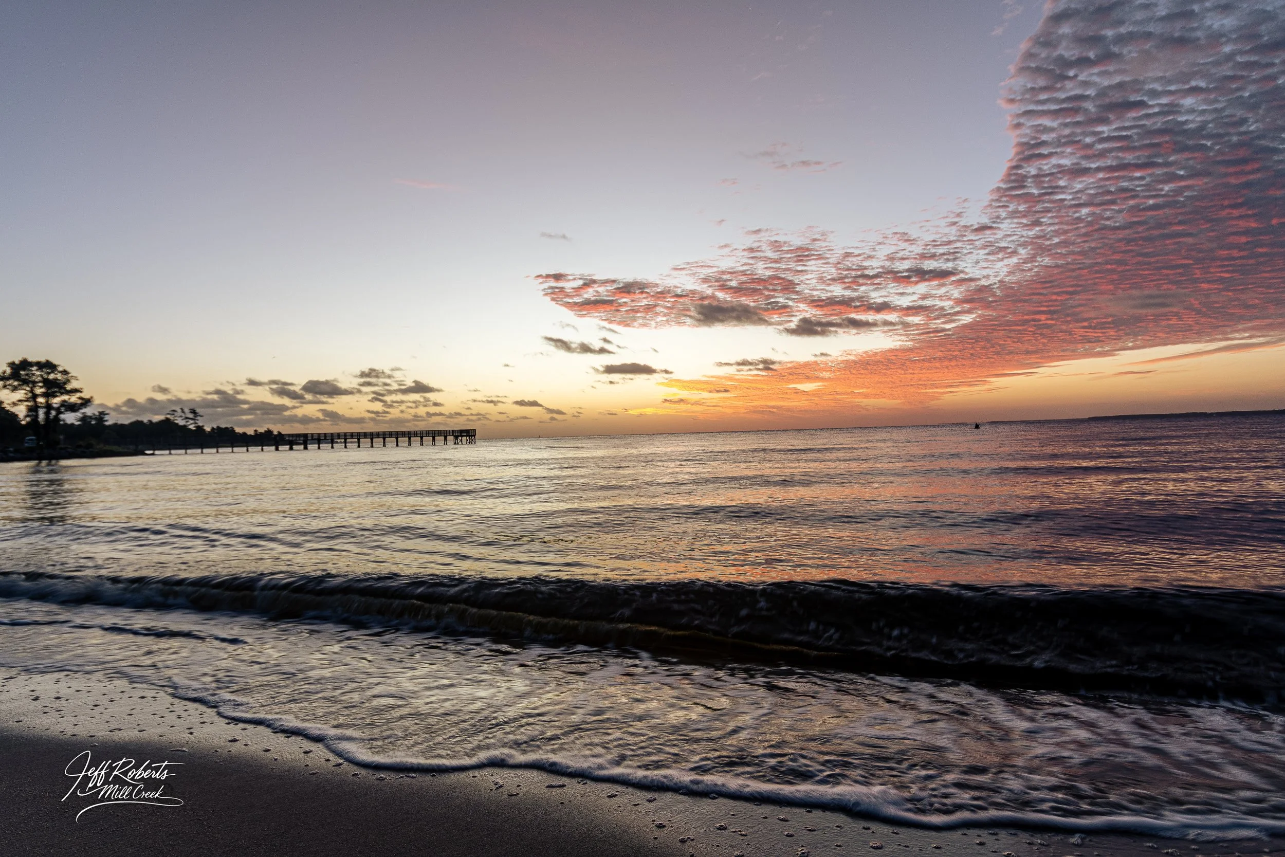Sunset over the ocean with a pier on the left side and a partly cloudy sky with pink and orange hues