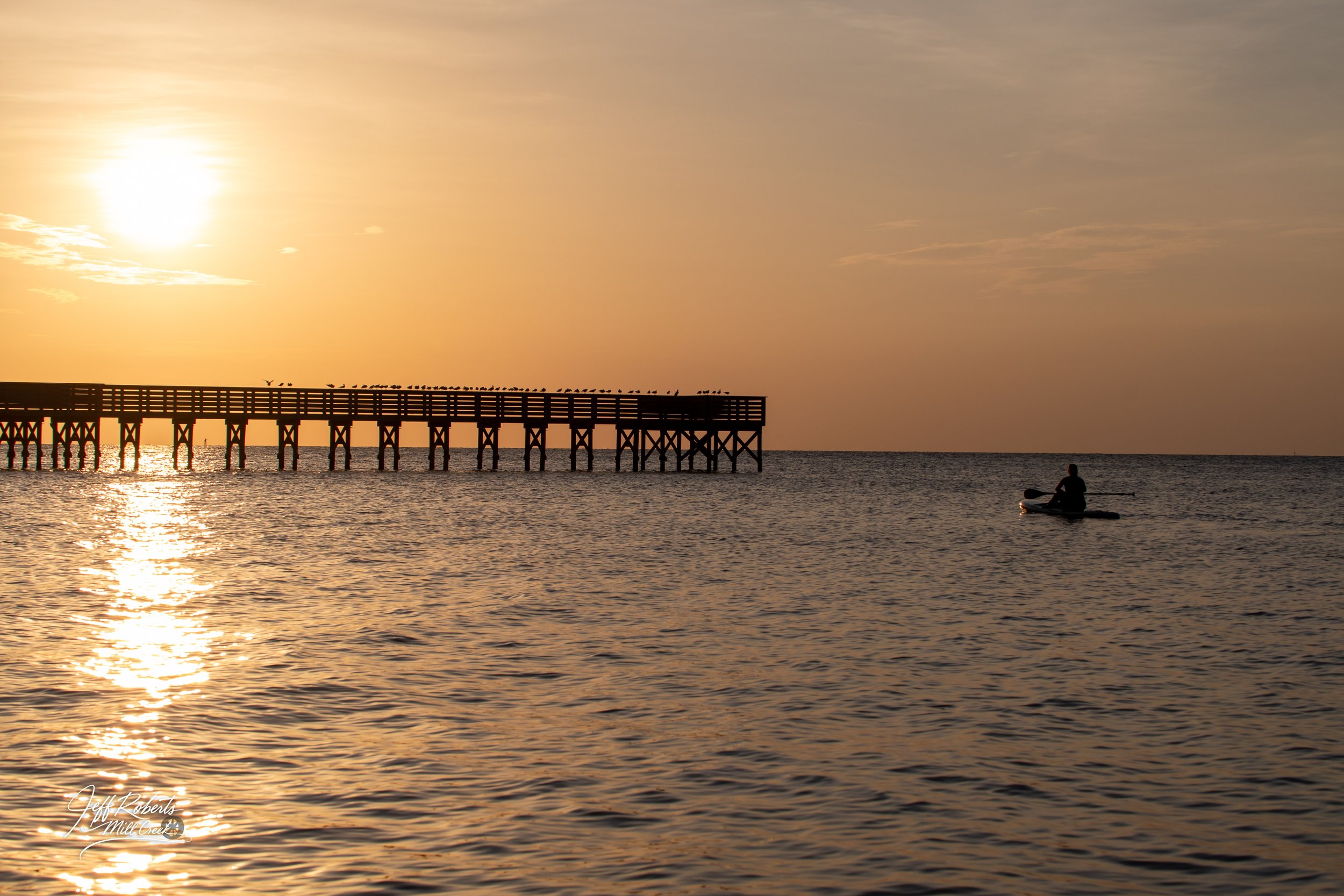 A person paddleboarding on calm ocean waters during sunset with a pier extending into the water on the left side of the image.