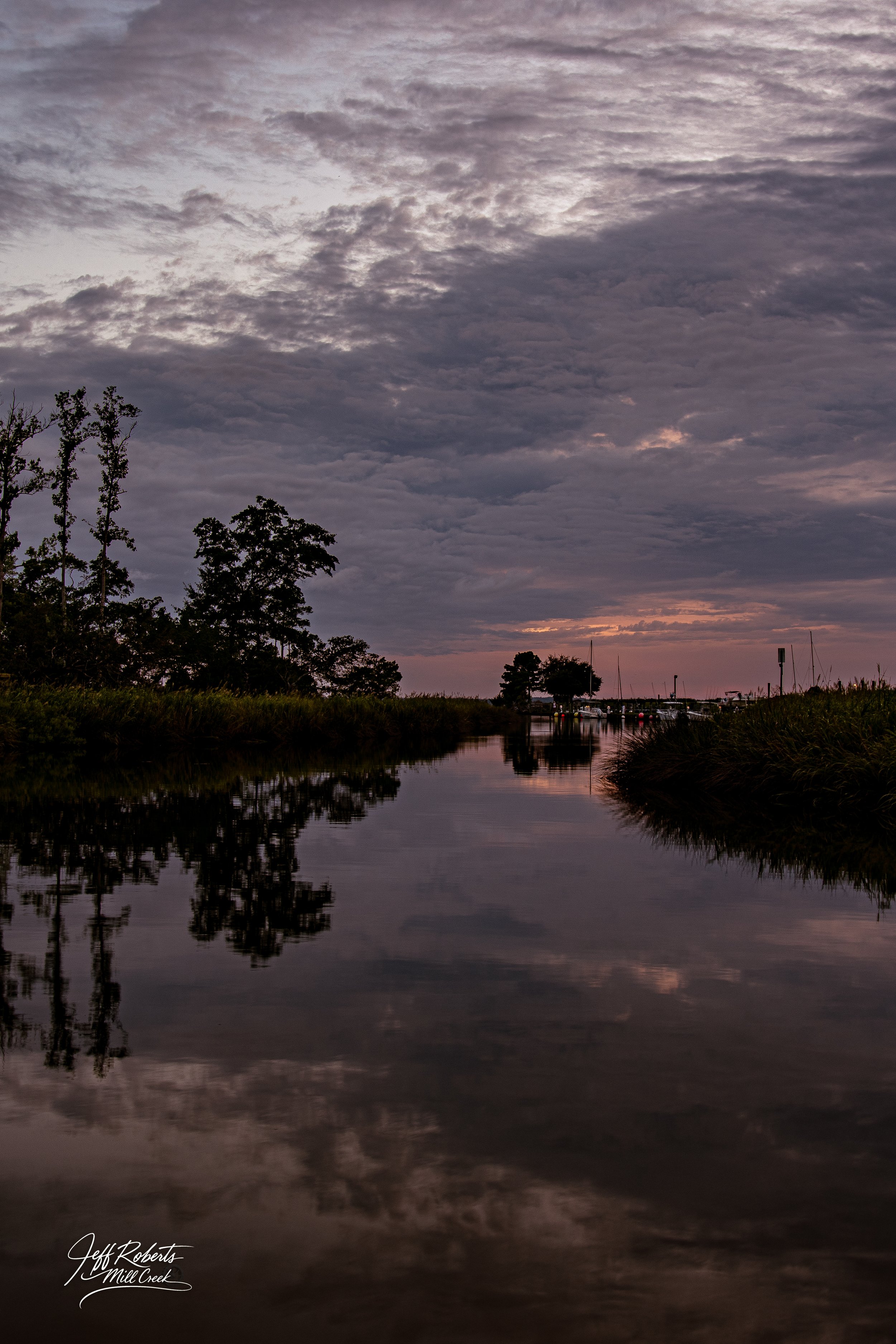 Sunset over a peaceful waterway with reflections of trees and clouds, and boats docked in the distance.