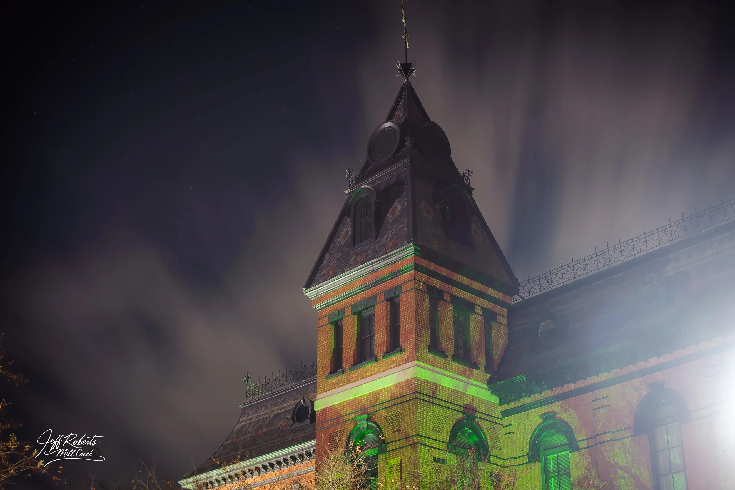 Night view of a historic brick building with a steep roof and tower, illuminated by colorful lights, with a cloudy sky in the background.