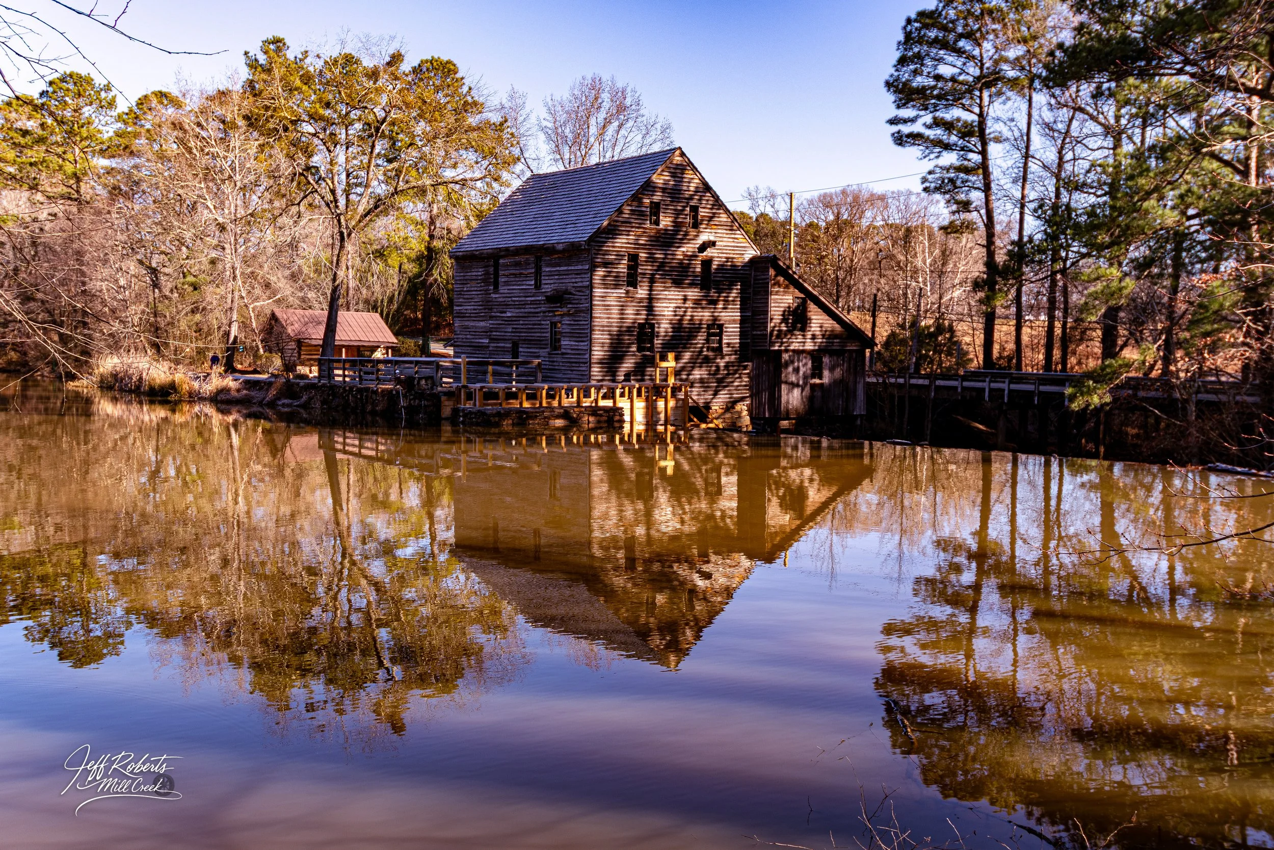 Old wooden mill house reflected in a calm river, surrounded by trees with bare branches and some evergreen trees, under a clear blue sky.