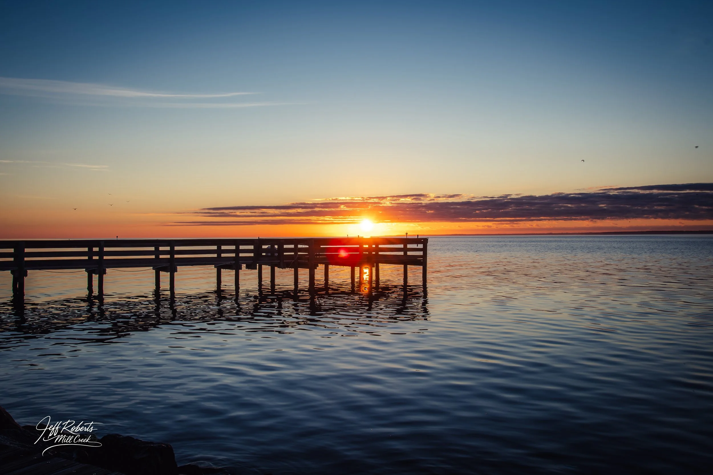 Sunset over a body of water with a wooden pier extending into the water, scattered birds in the sky, and clouds on the horizon.