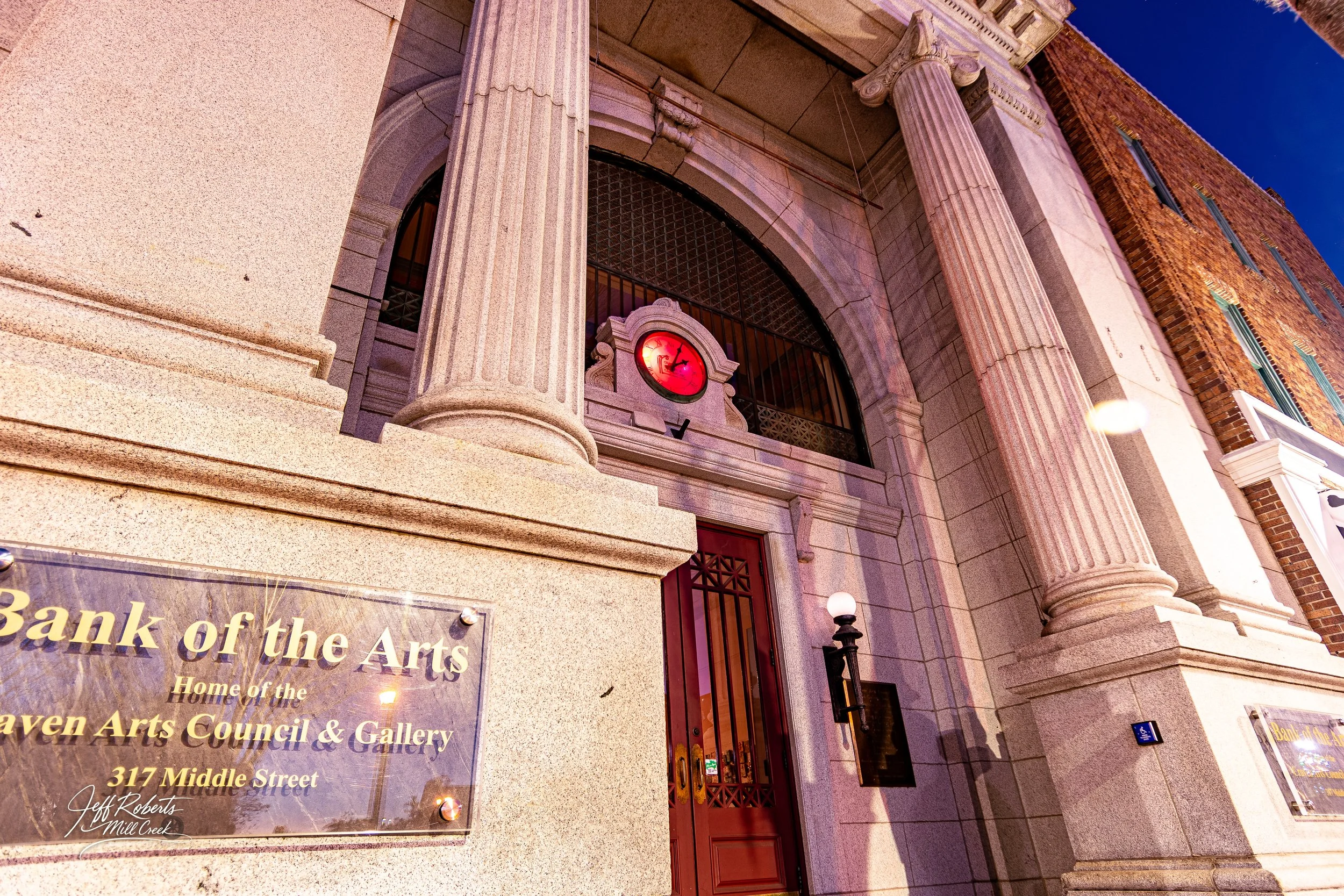 Front view of the Bank of the Arts building with large stone columns, an arched window, and a red clock above the entrance at sunset.