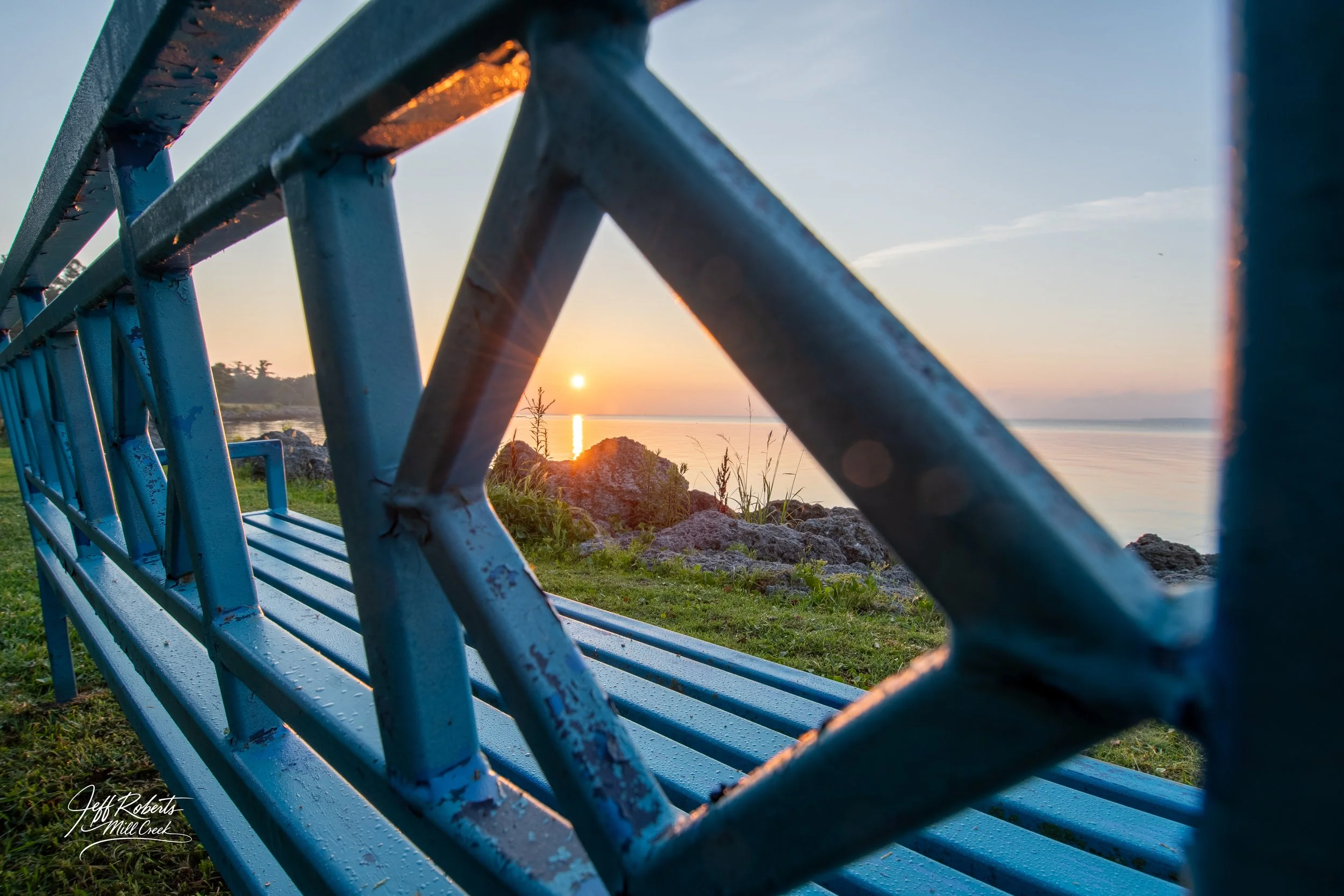 View of a sunset over a body of water, seen through a blue bench with a diamond-shaped opening in an outdoor setting with rocks and grass