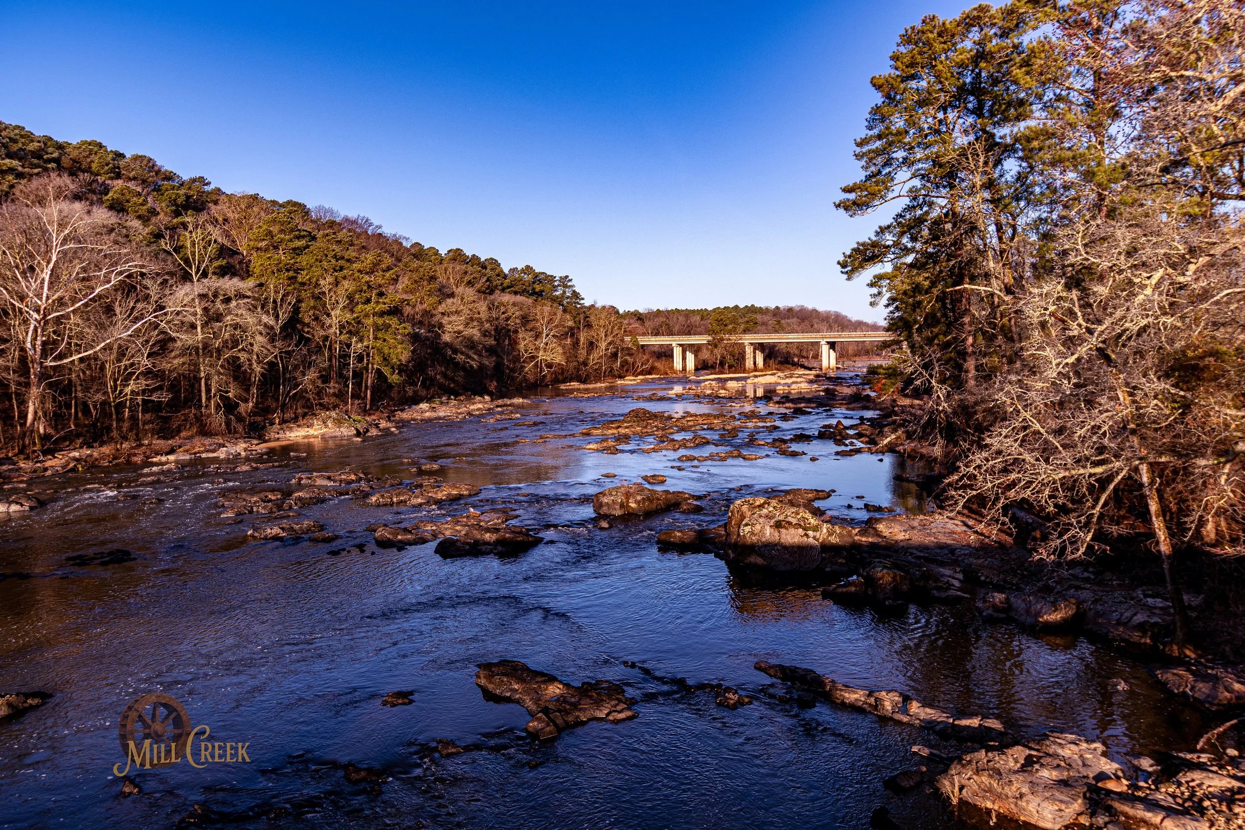 Scenic view of a river with rocks, surrounded by trees, some with bare branches, and a bridge in the background.