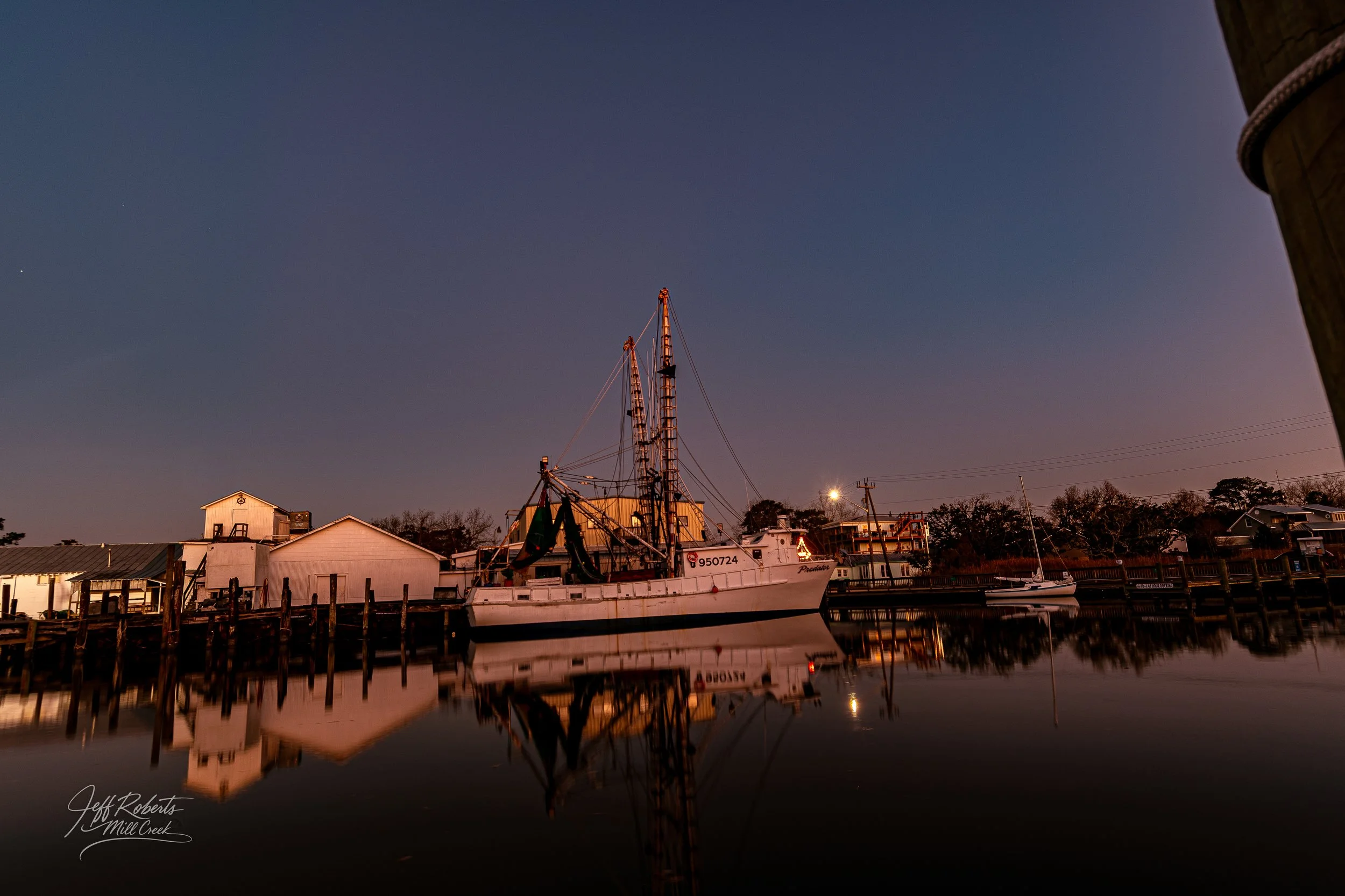 A boat docked along the water at dusk with houses and trees in the background, and the boat's reflection visible in the water.