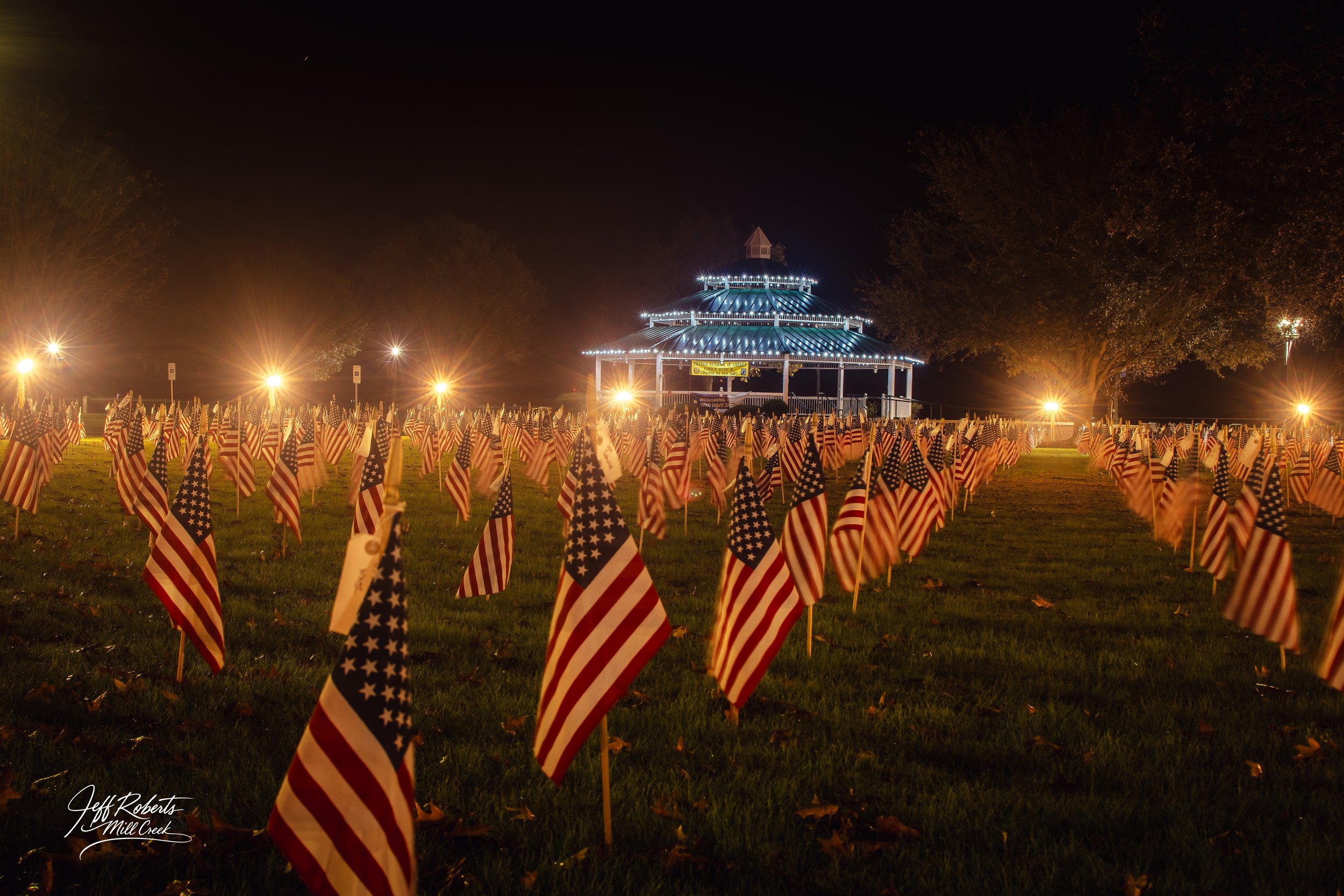 Nighttime outdoor scene with numerous small American flags planted in grass, leading to a brightly lit, multi-tiered pavilion in the background decorated with white string lights, and tall trees on sides.