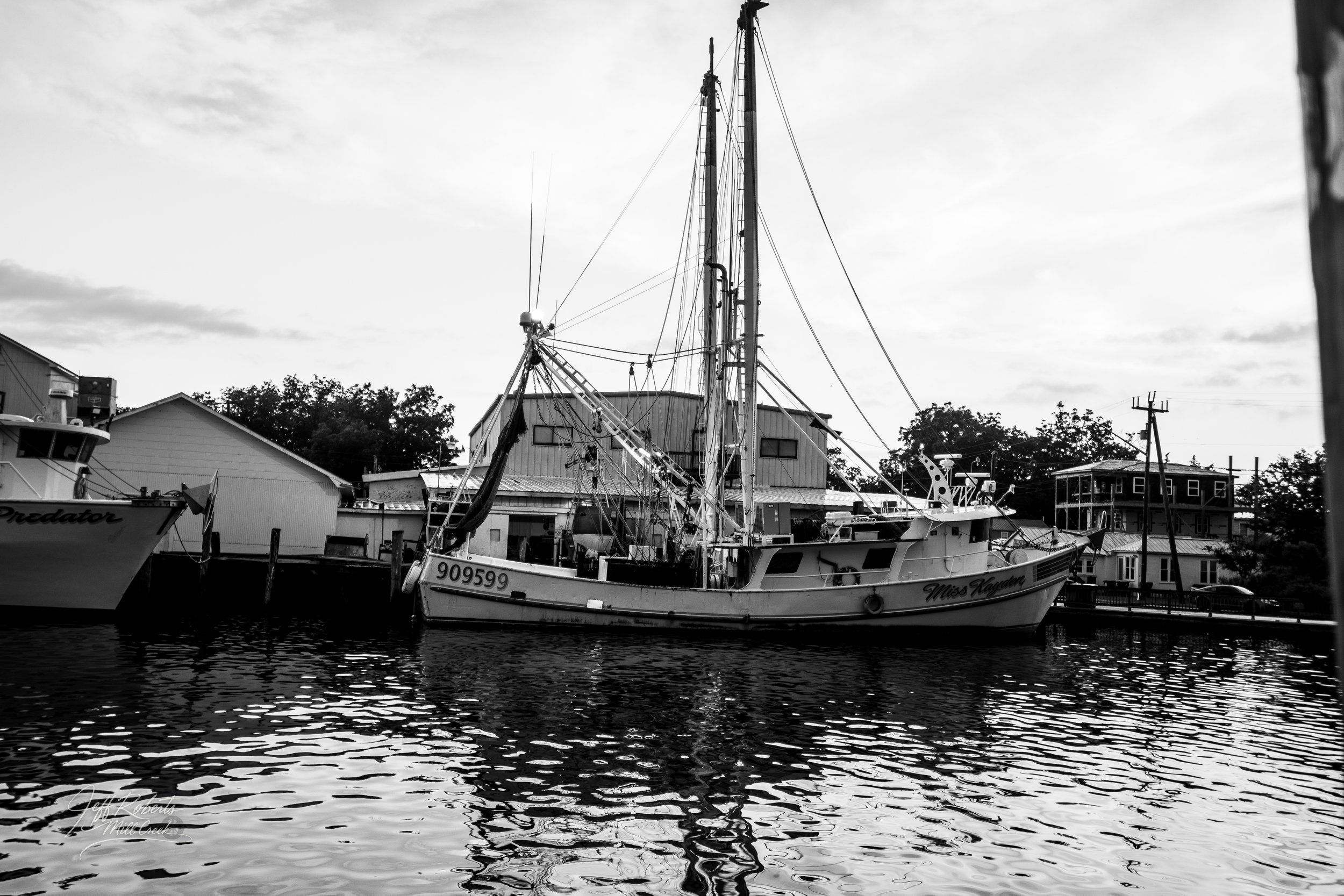 A black and white photo of a fishing boat named Miss Kayden docked in a marina with buildings and trees in the background.