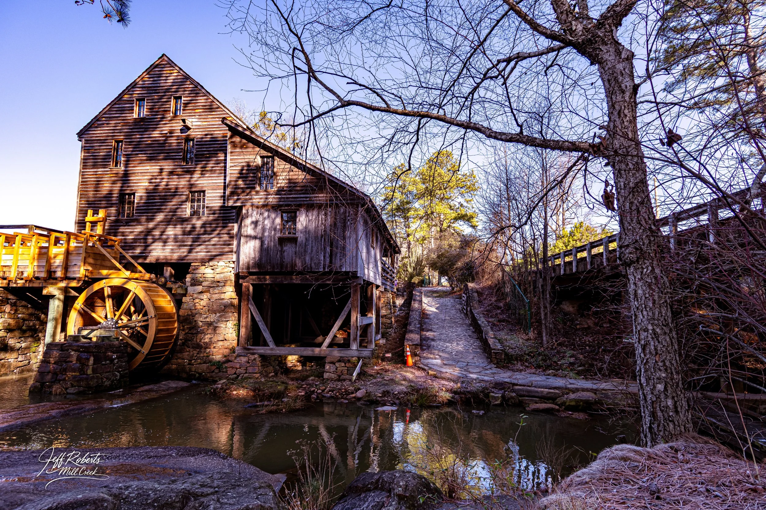 An old wooden mill with a water wheel next to a small creek, surrounded by leafless trees, with a stone pathway leading up a hill in the background.