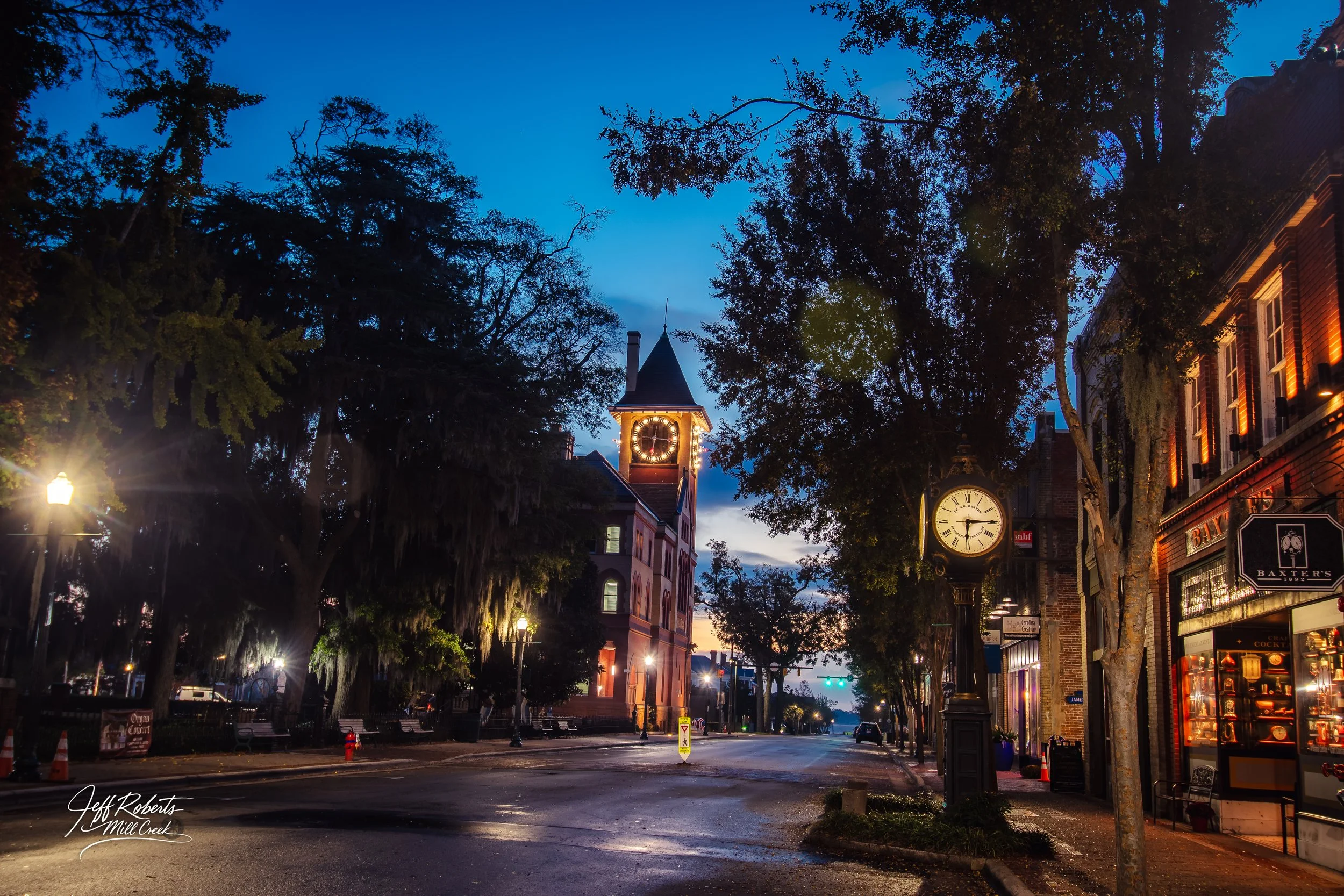 City street at dusk with a clock tower, street lamps, trees, and storefronts illuminated.