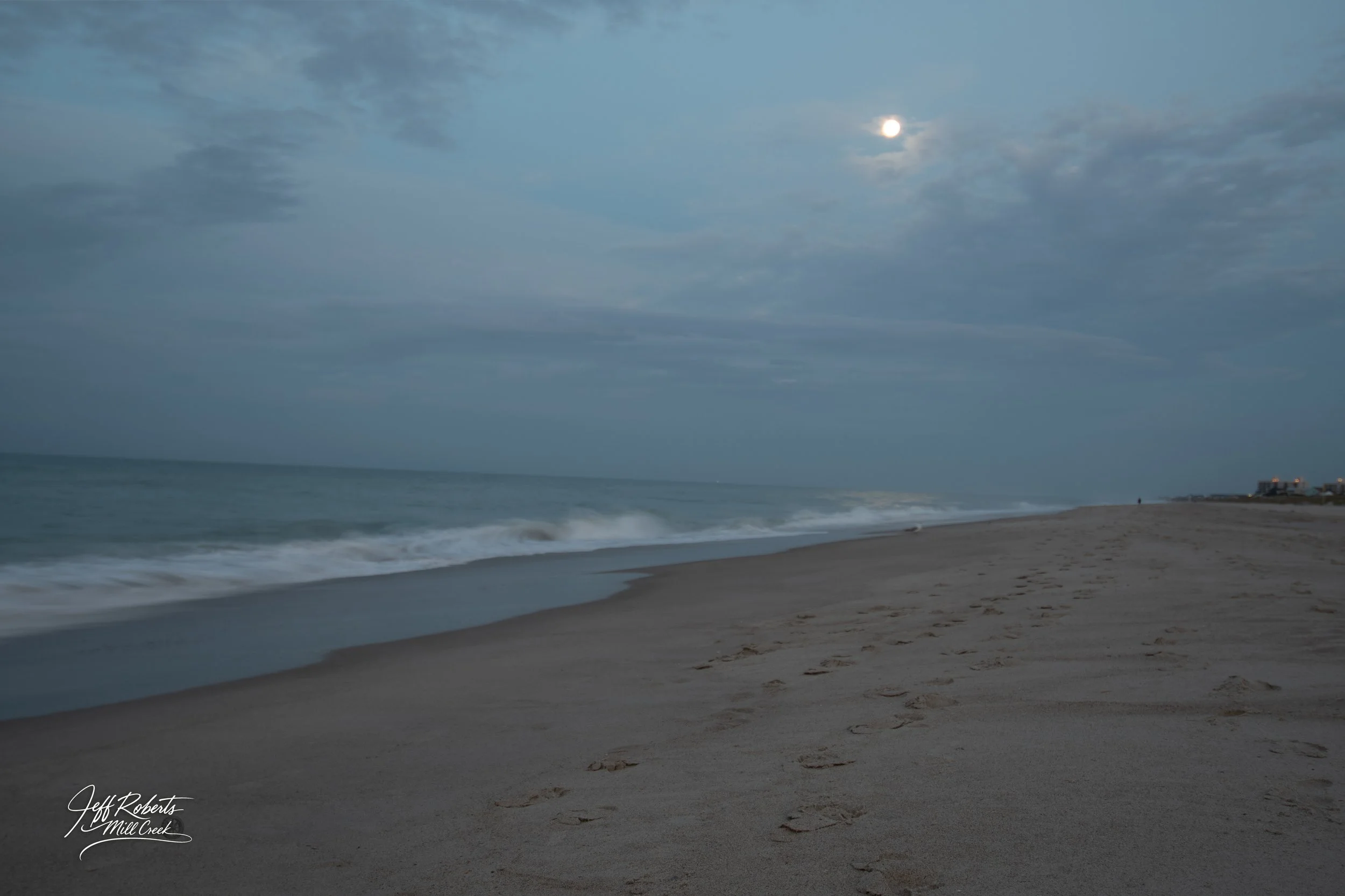 A peaceful beach scene under a cloudy sky with the moon visible, gentle waves lapping the shore, and footprints in the sand.