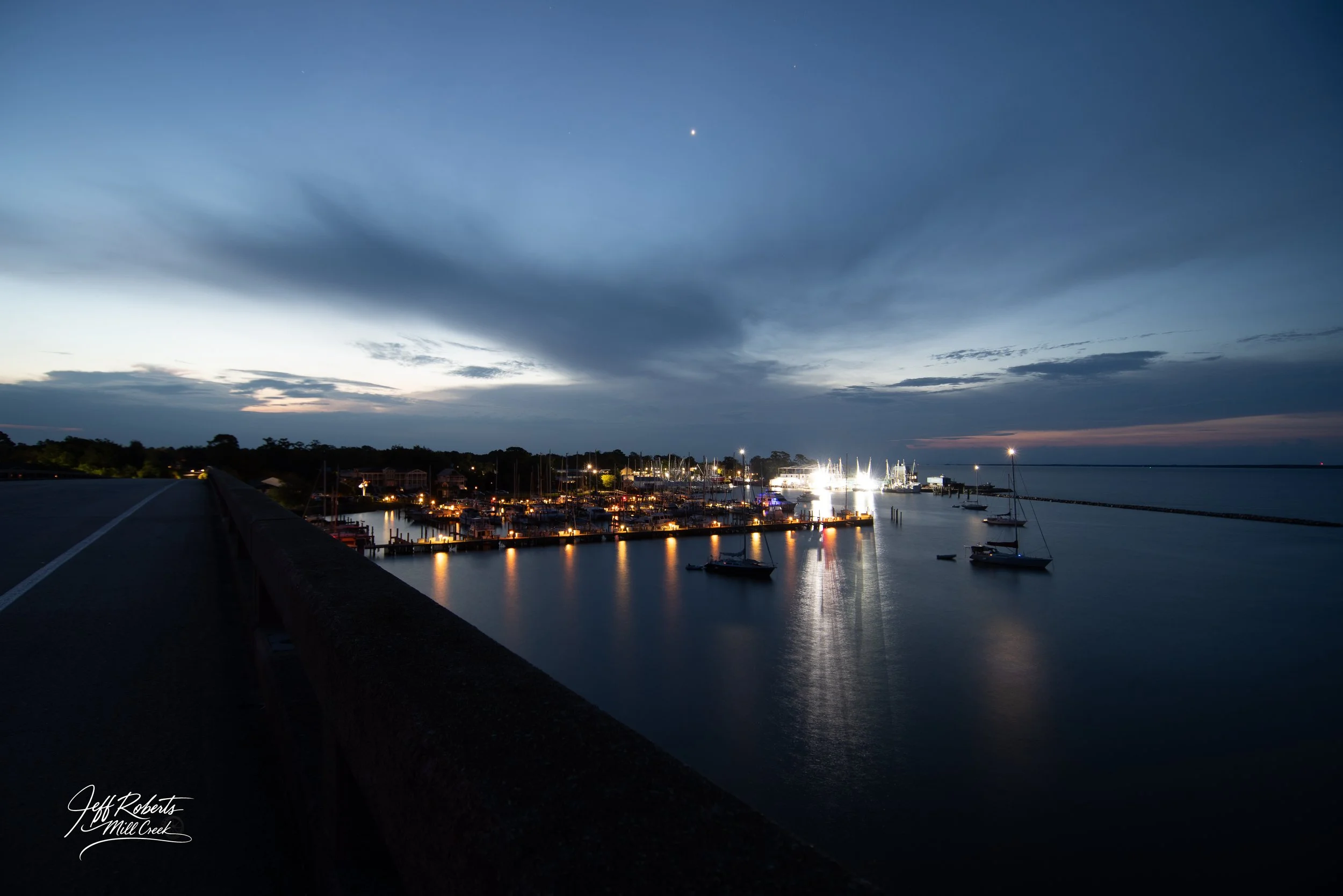 Night view of a marina with boats docked, illuminated by lights, under a cloudy sky at dusk or early evening.