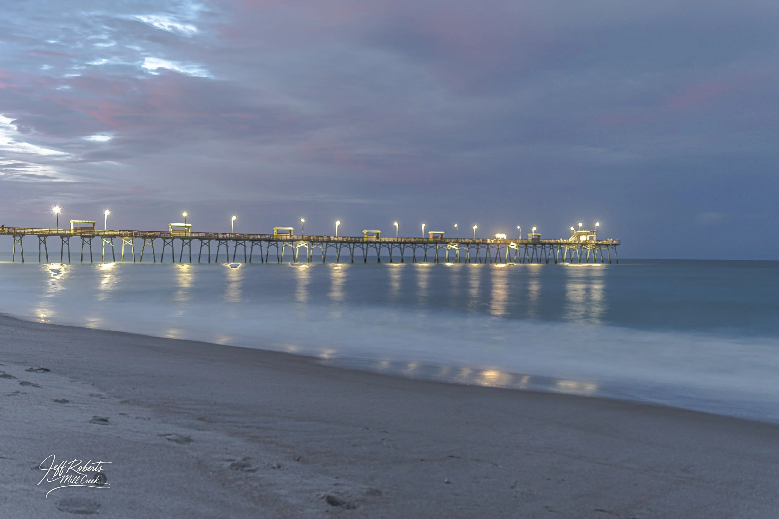Photo of a pier extending into the ocean at dusk, with lights illuminating the structure and reflecting on the water. The sky is partly cloudy with a mix of blue and purple hues, and the sandy beach is visible in the foreground.