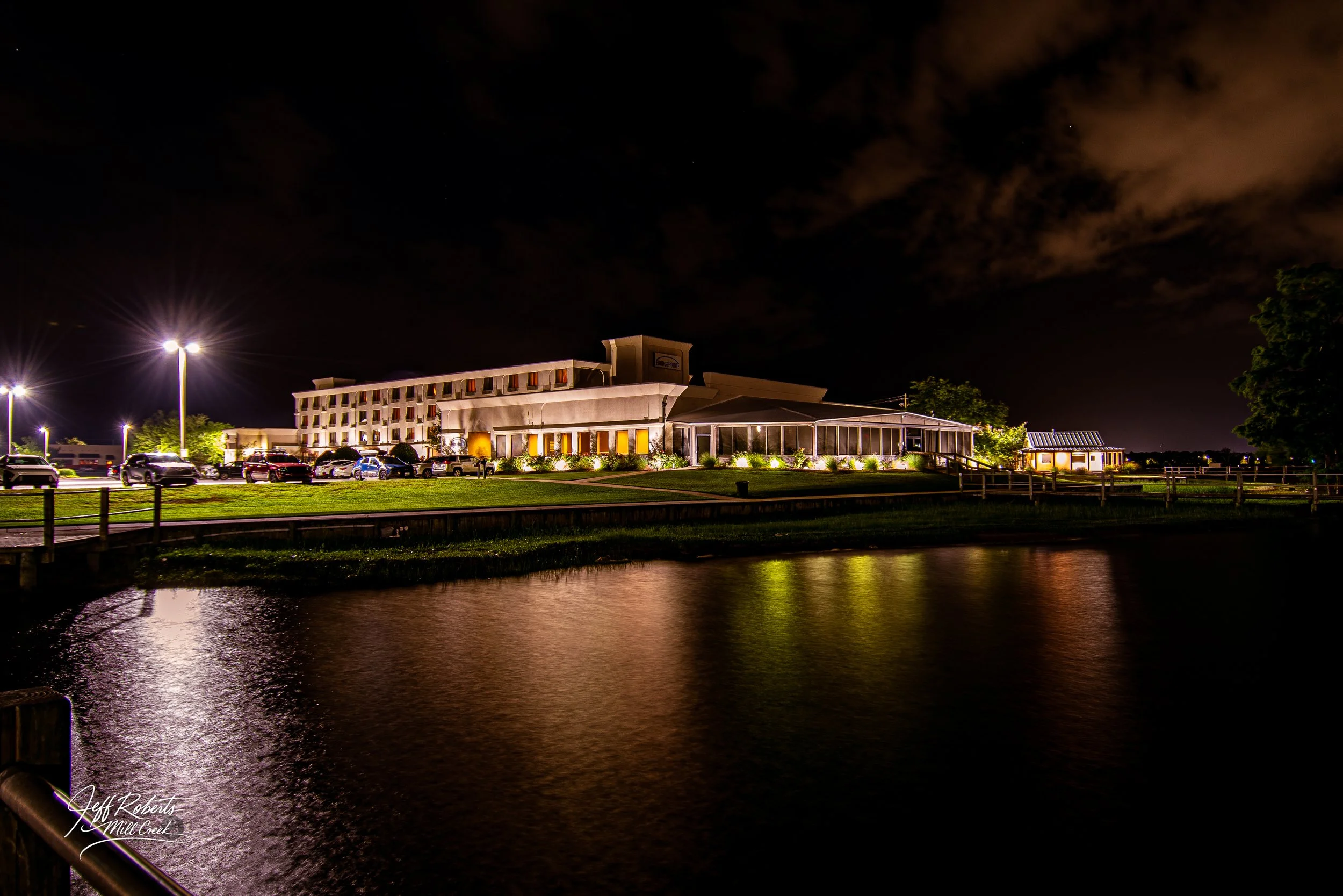 Nighttime view of a well-lit hotel building with multiple cars parked in front, surrounded by streetlights, trees, and a body of water reflecting the lights.