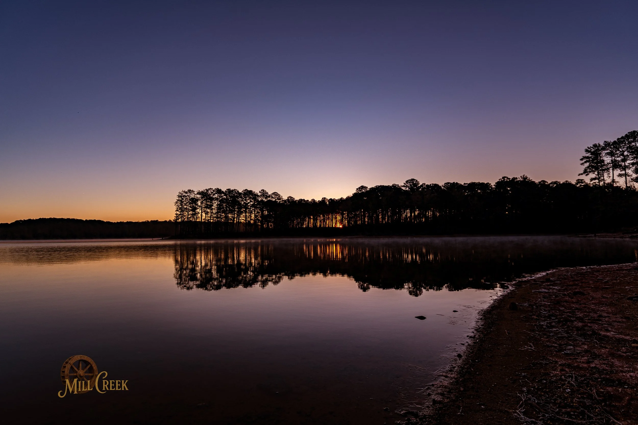 A calm lake at sunset with a silhouette of trees on the horizon and a reflection on the water. Mill Creek logo in the bottom left corner.