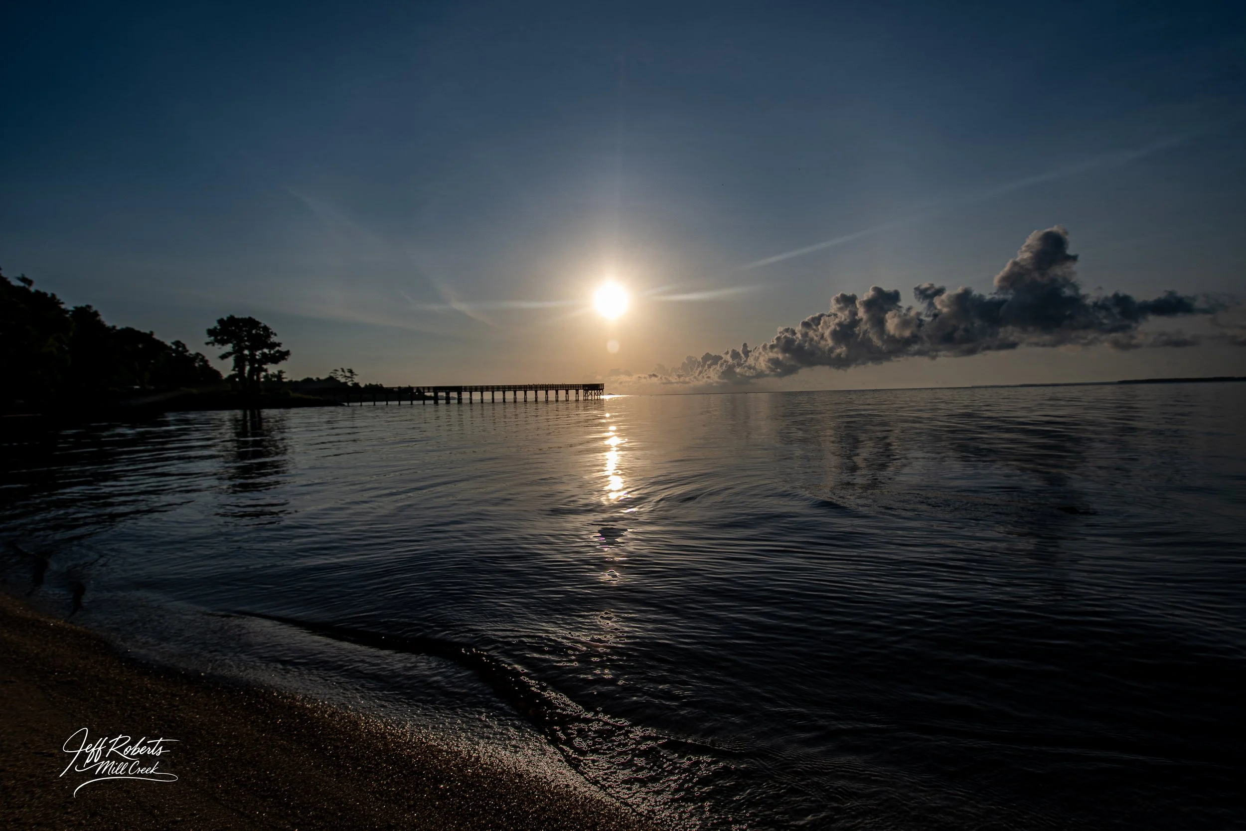 Sunset over a calm body of water with a wooden pier extending into the distance, silhouetted trees on the left, and clouds on the horizon.
