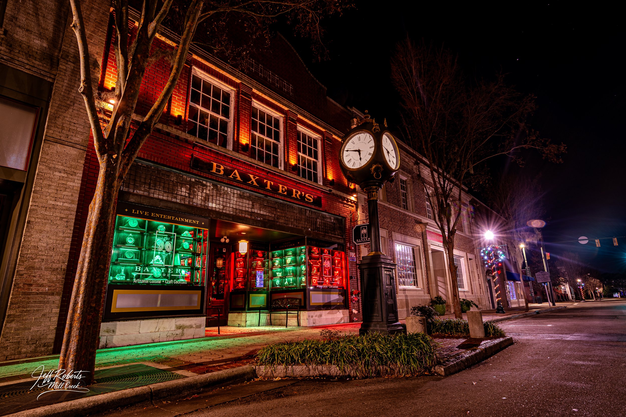 Night street scene featuring Baxtor's bar with colorful neon lights, a large clock on a street pole, leafless trees, and a few illuminated buildings in the background.