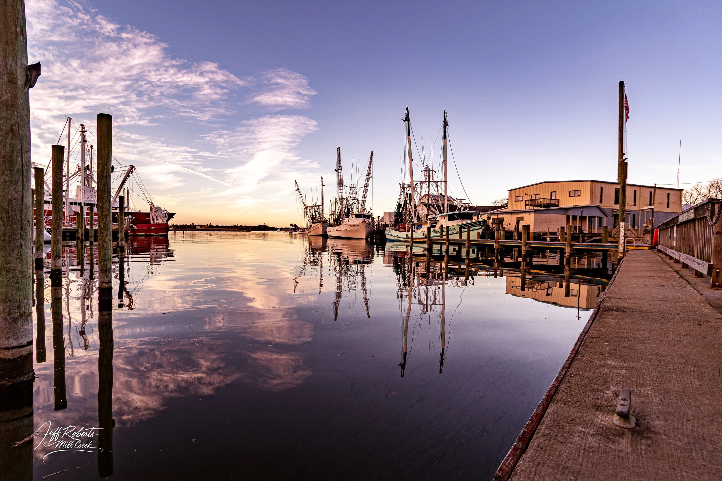 Dock at a marina with boats reflecting in calm water during sunset, with a building and a wooden pier on the right side.