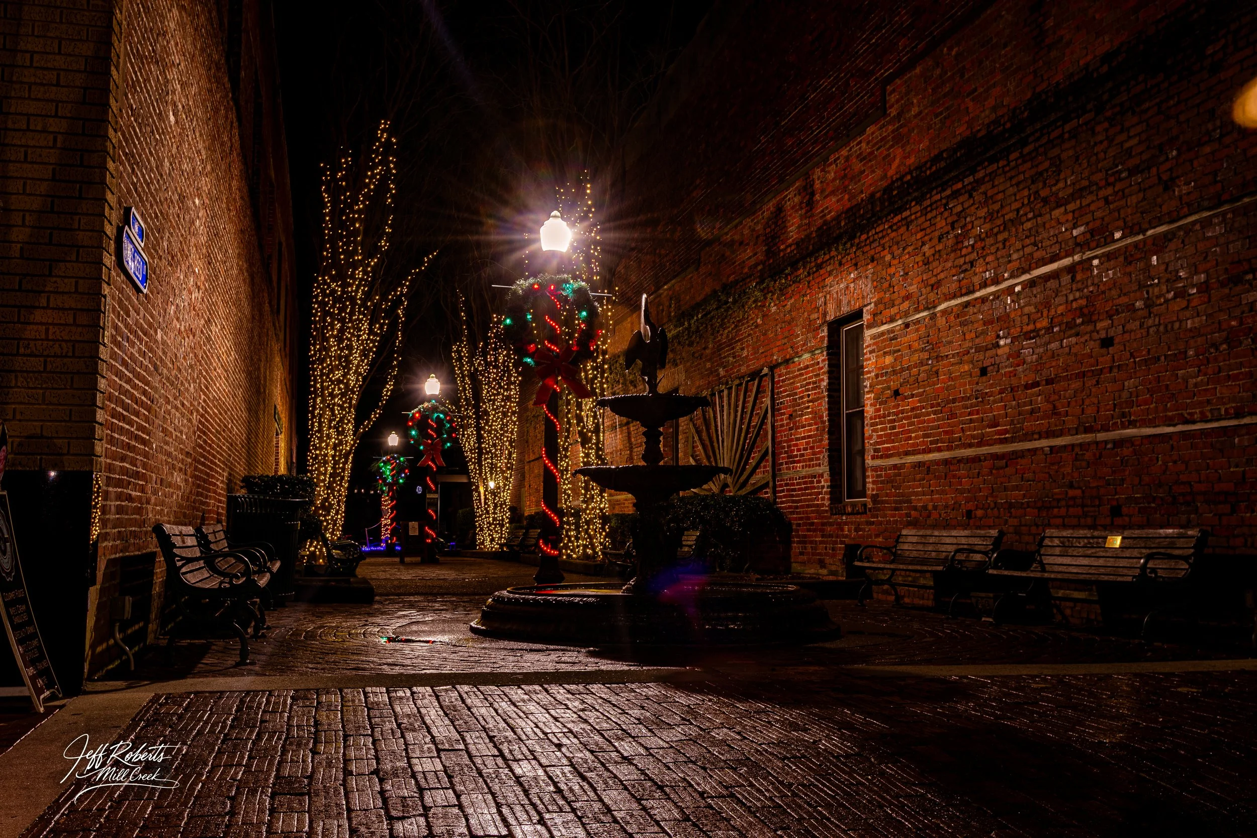 Nighttime view of a decorated brick-lined alley with Christmas lights, wreaths, and benches.