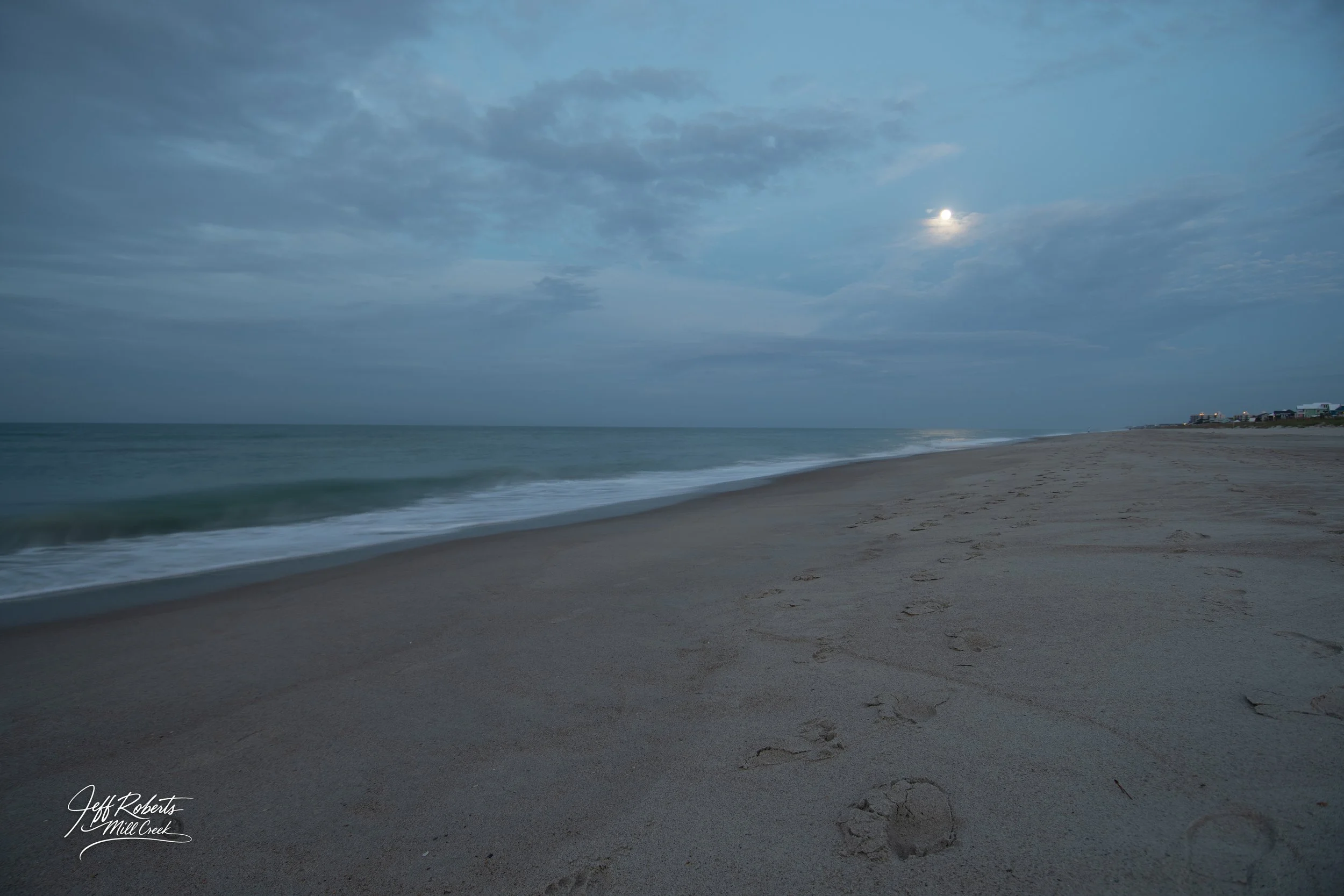 A quiet beach scene at dusk with footprints in the sand, calm ocean waves, partly cloudy sky, and the moon visible.