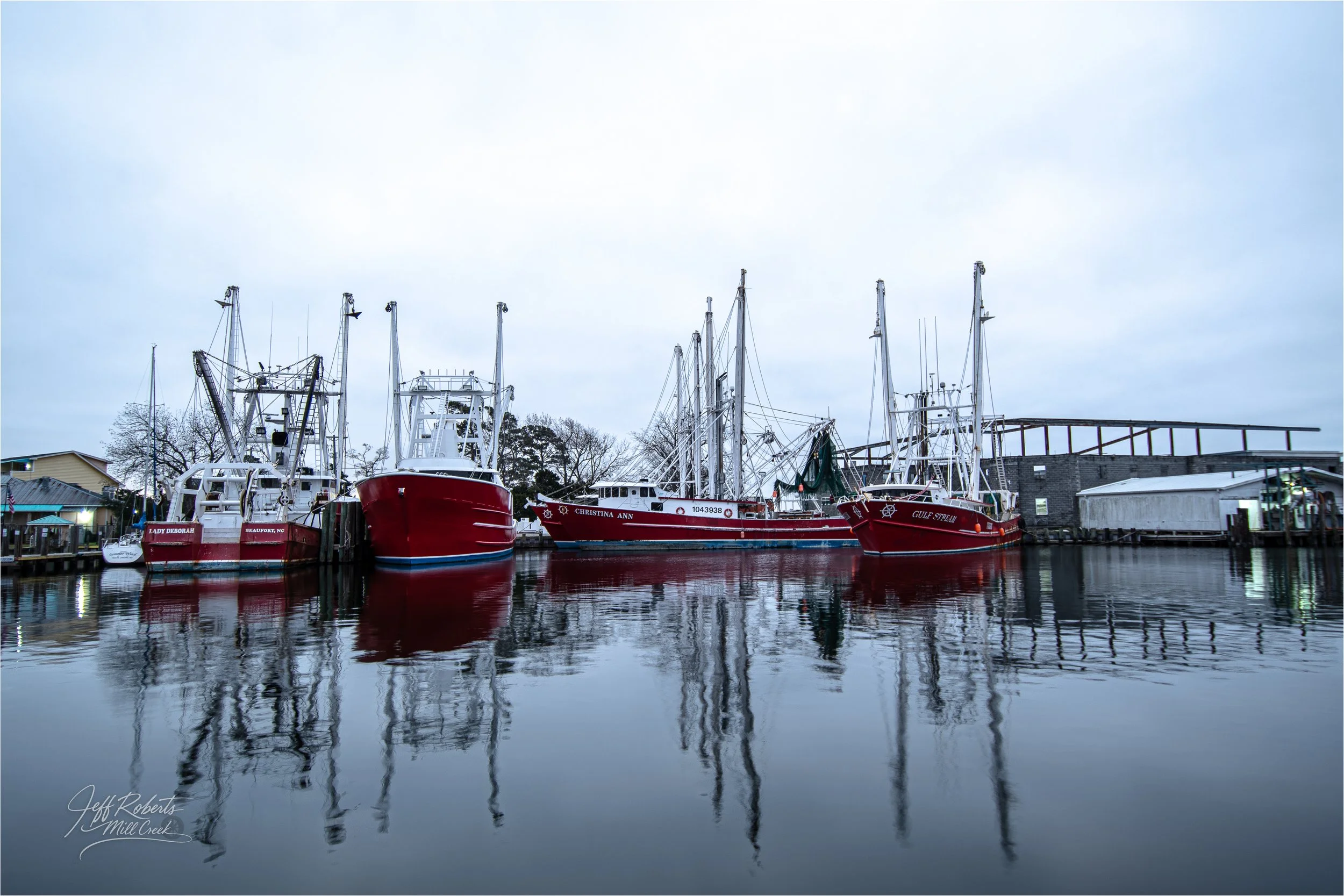 Four red fishing boats docked at a marina with calm water reflecting the boats, under an overcast sky.