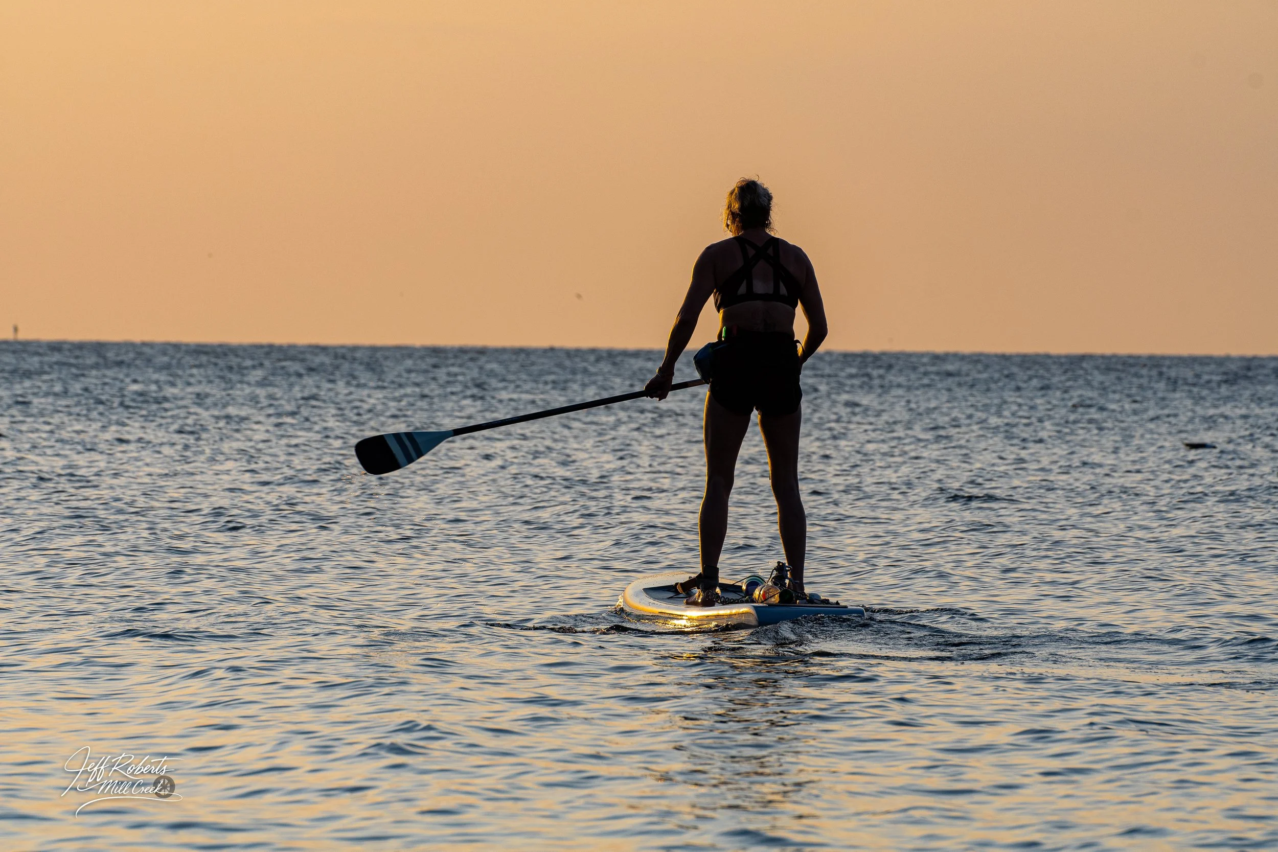 Silhouette of a woman paddleboarding on the water during sunset with a calm ocean and orange sky in the background.