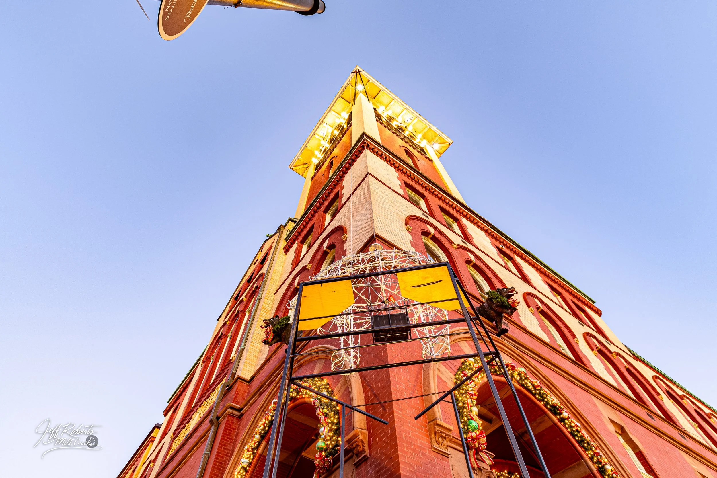 Low-angle view of a tall, decorated red brick building with Christmas ornaments and lights, topped with a yellow roof, against a clear blue sky at dusk.