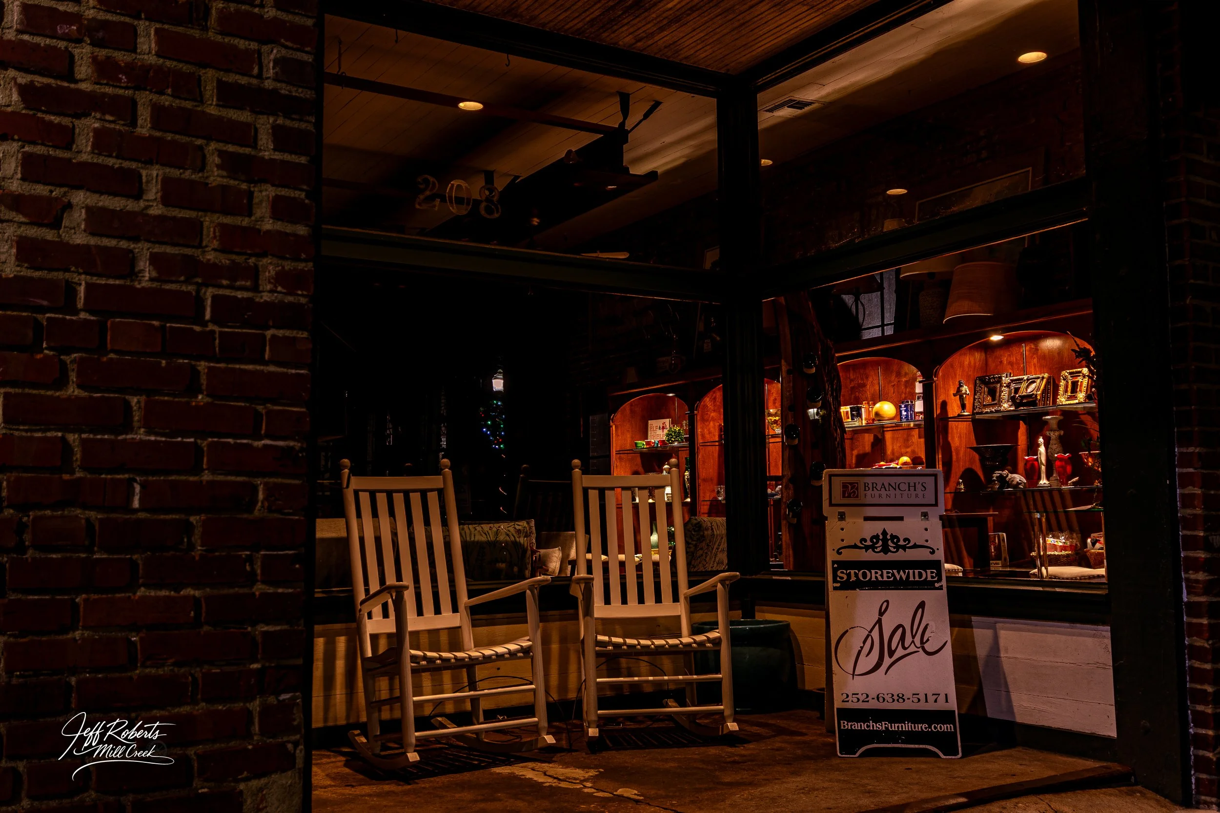 The image shows the front of a furniture store at night with two wooden rocking chairs in front of a large window. Inside, decorated shelves with various items and a Christmas tree in the background are visible, creating a cozy, festive atmosphere.