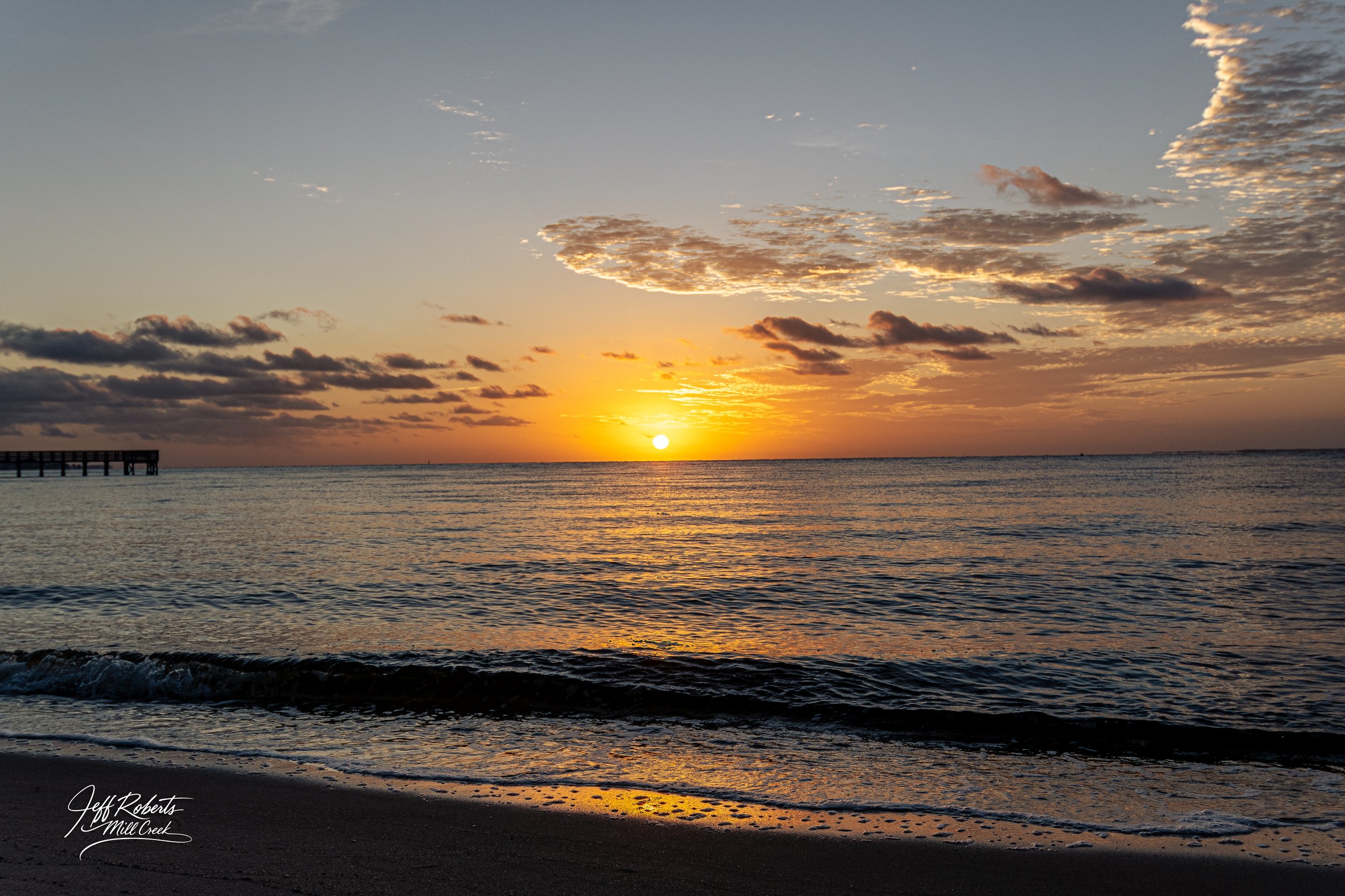 Sunset over the ocean with a sandy beach in the foreground and a pier on the left