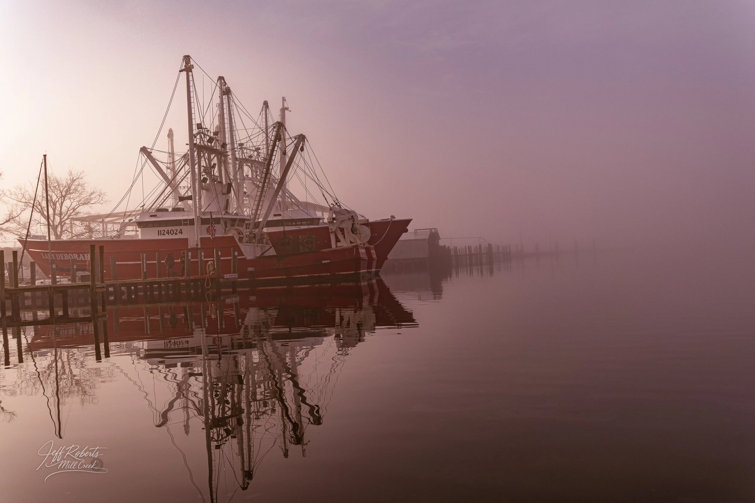 A red fishing boat named 'Lyn Deborah' docked at a pier on a calm, foggy morning, with reflections seen in the still water.